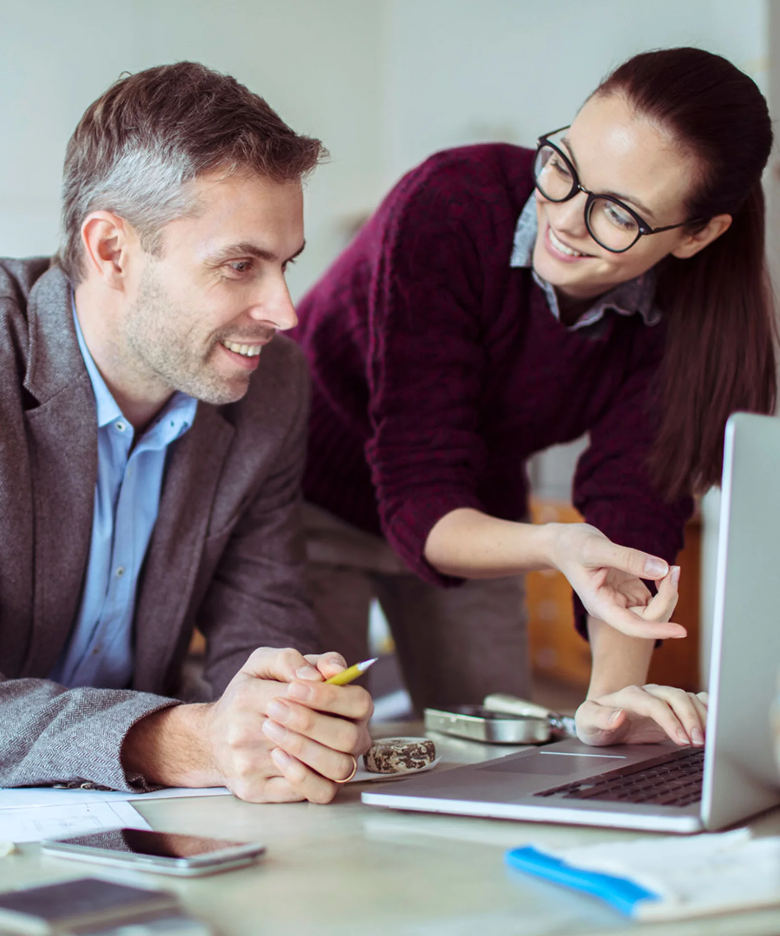 Two colleagues collaborating on a project, with a woman pointing at a laptop screen and a man smiling, seated at a desk in a modern office.