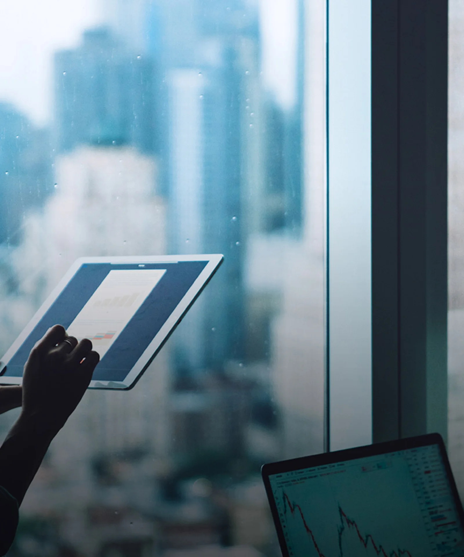 Woman analyzing financial data on a tablet while standing near a window with a cityscape background, showcasing digital transformation in business environments.