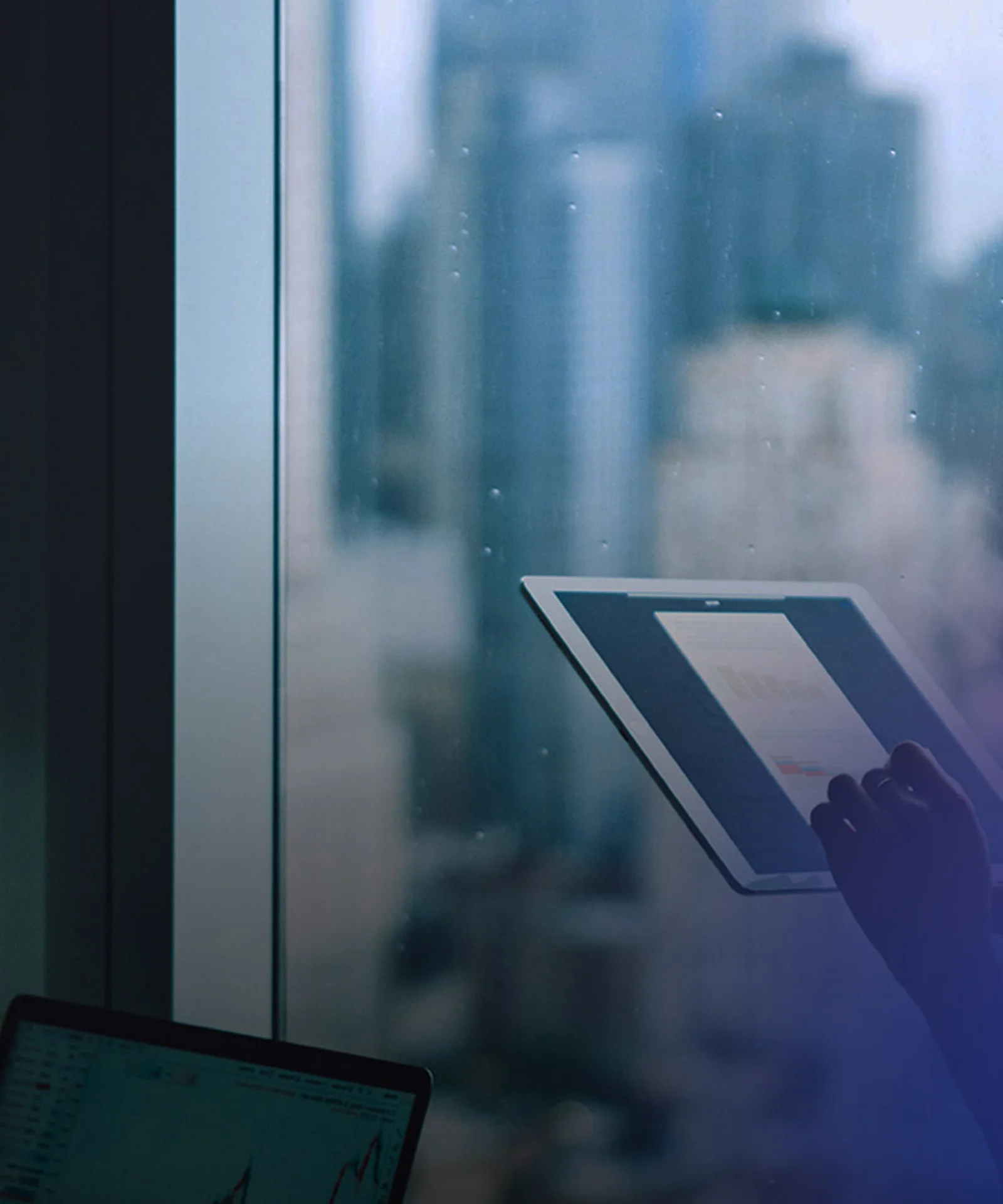 A businesswoman standing by a window, using a tablet to analyze data, with city skyscrapers blurred in the background.