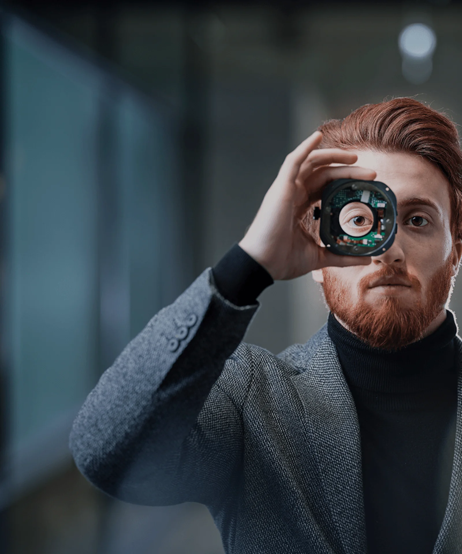 A man with red hair and beard holds a technological component in front of his eye, symbolizing innovation and focus. He is dressed in a grey suit and stands in a modern, dimly lit environment.