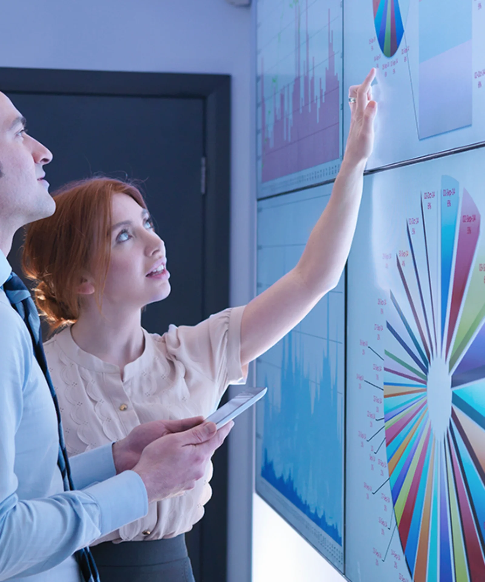 Two business analysts examining large digital screens displaying complex data visualizations, graphs, and charts, focused on analyzing trends.