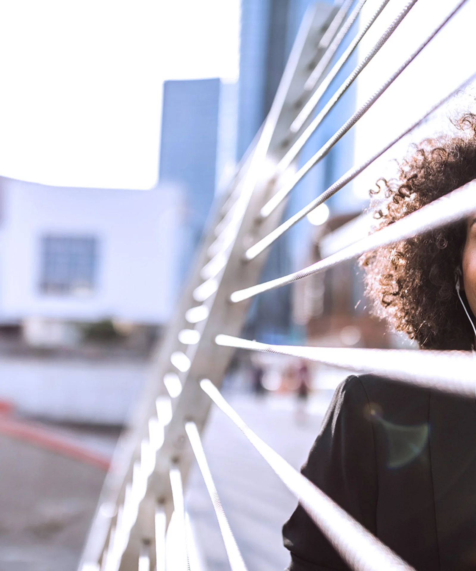 Smiling professional woman with curly hair, listening to music on a modern bridge in an urban setting.