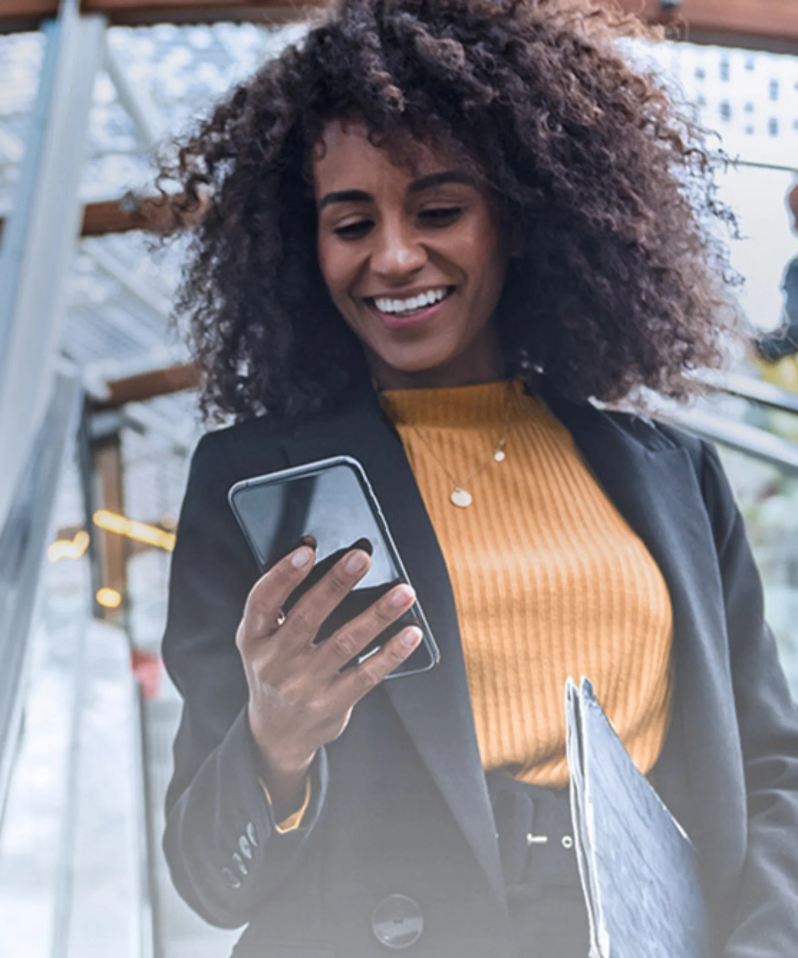 Smiling businesswoman using a smartphone while riding an escalator in a modern building with glass and metal architecture.