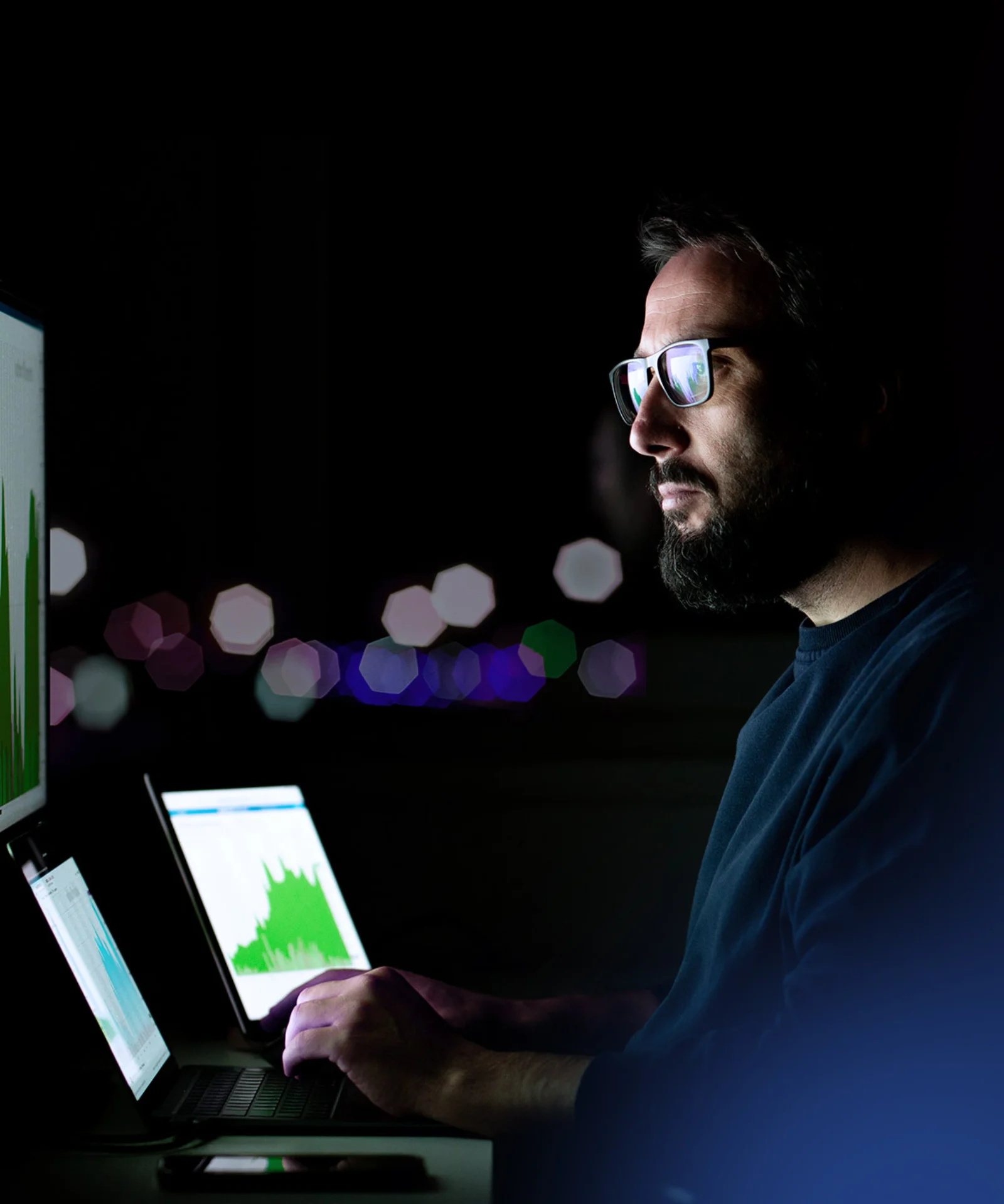 A man analyzing data on multiple screens in a dark room, focused on charts and graphs, with soft, blurred lights in the background.