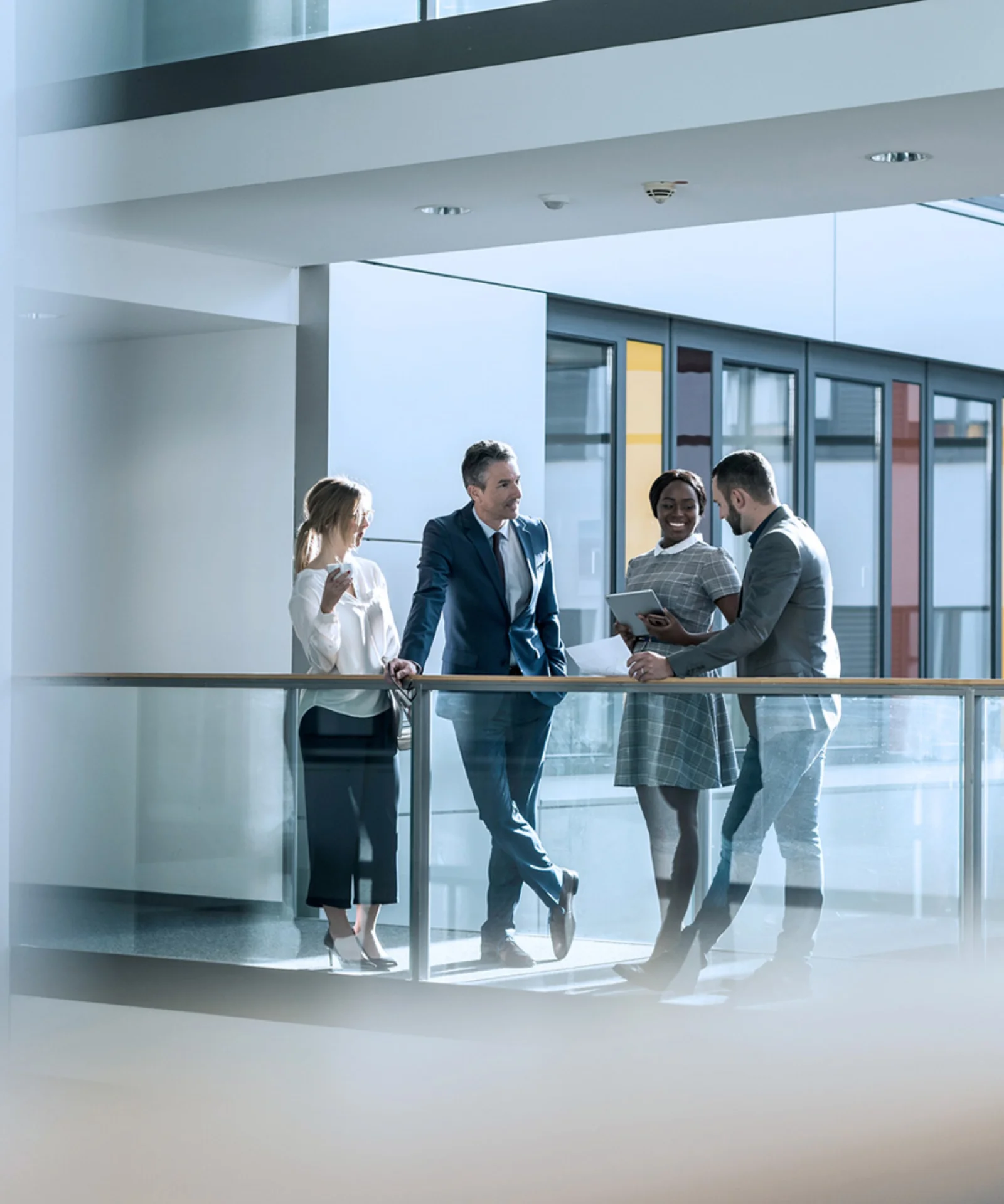 This image captures a professional meeting in a modern office setting. Four individuals, three men and one woman, are engaged in a conversation, standing by a glass railing on an upper floor. The office environment features clean, contemporary design elements with large windows and glass partitions, creating a bright and open atmosphere. The people are dressed in business attire, suggesting a formal and collaborative business environment. This visual is ideal for conveying partnership, collaboration, and a modern business ethos.