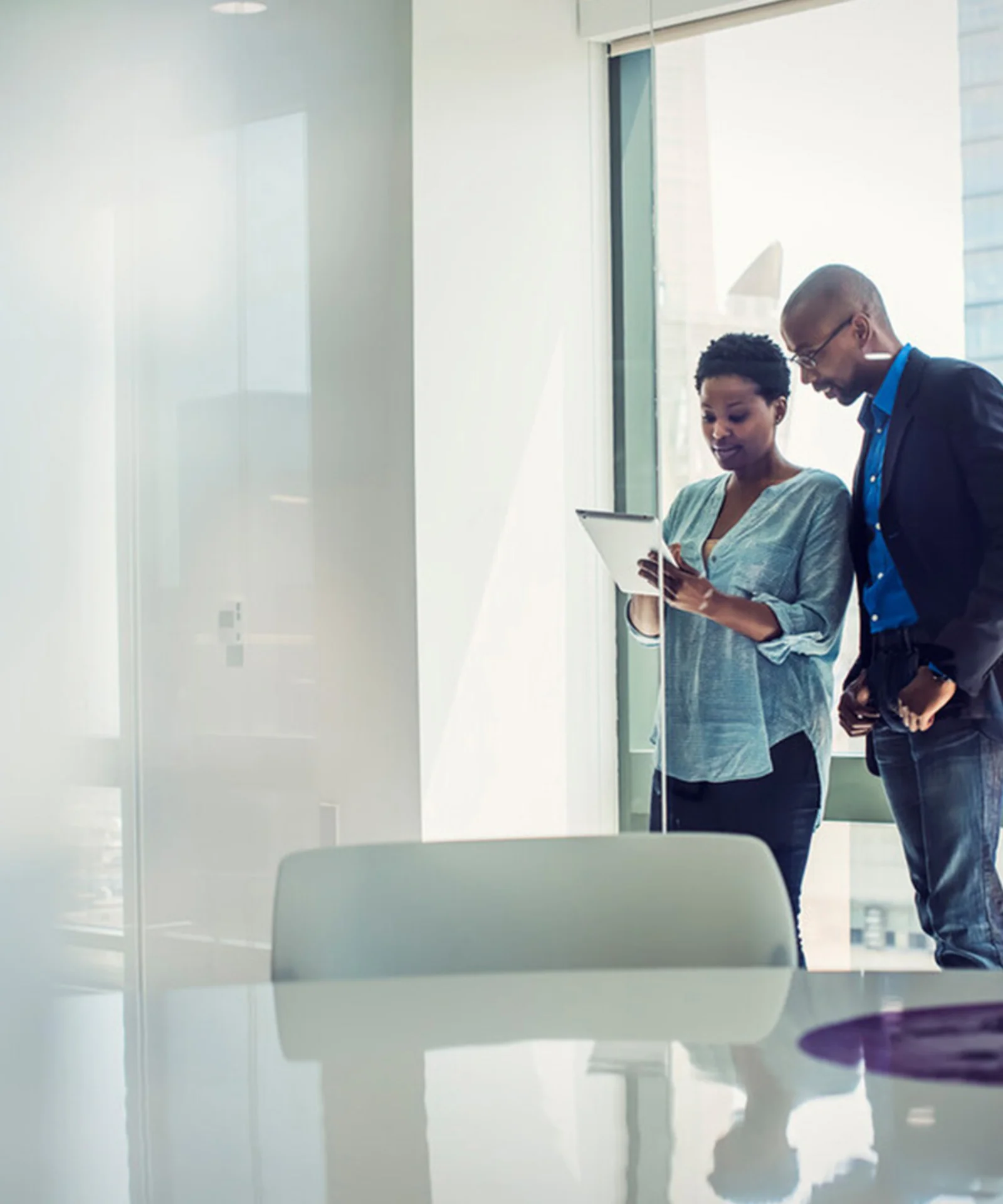 Two business professionals reviewing information on a tablet in a modern, bright office with large windows.