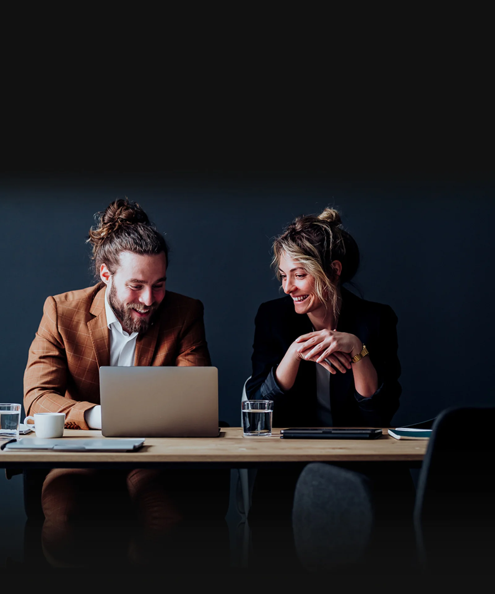 Two professionals engaged in a collaborative discussion, sitting at a desk with a laptop, notebooks, and coffee, showcasing an image design services setup.