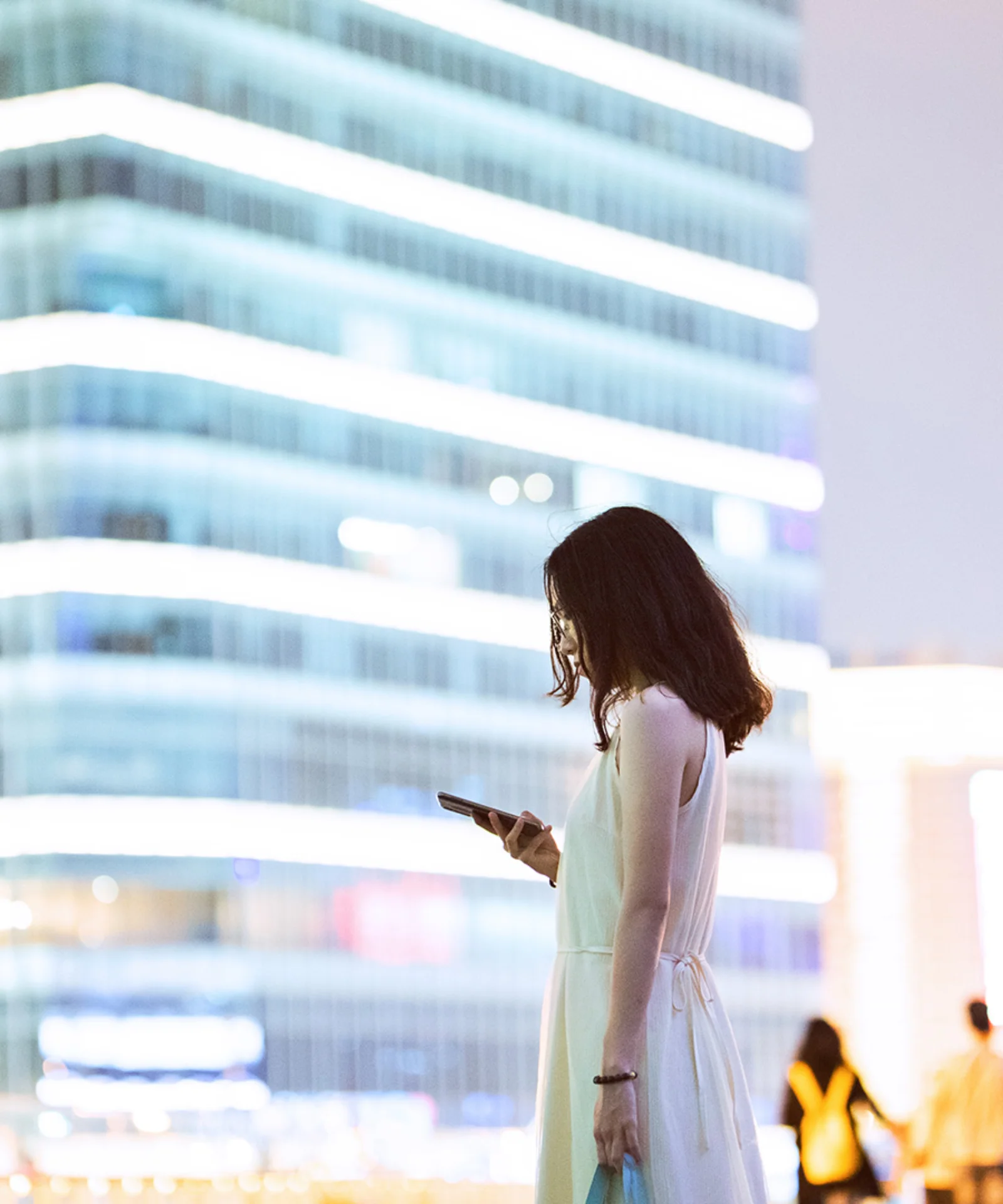 Une femme debout dans un cadre urbain la nuit, regardant son smartphone, avec des bâtiments modernes bien éclairés en arrière-plan, représentant le thème de la numérisation dans le secteur bancaire.