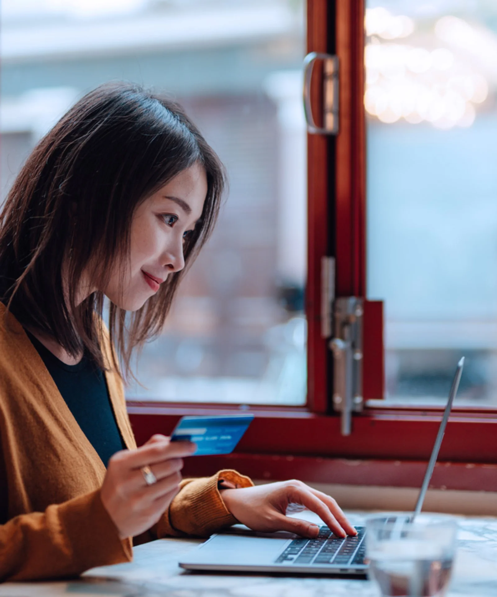 A woman holding a credit card while using a laptop in a cosy café setting, symbolising digital banking and convenience.