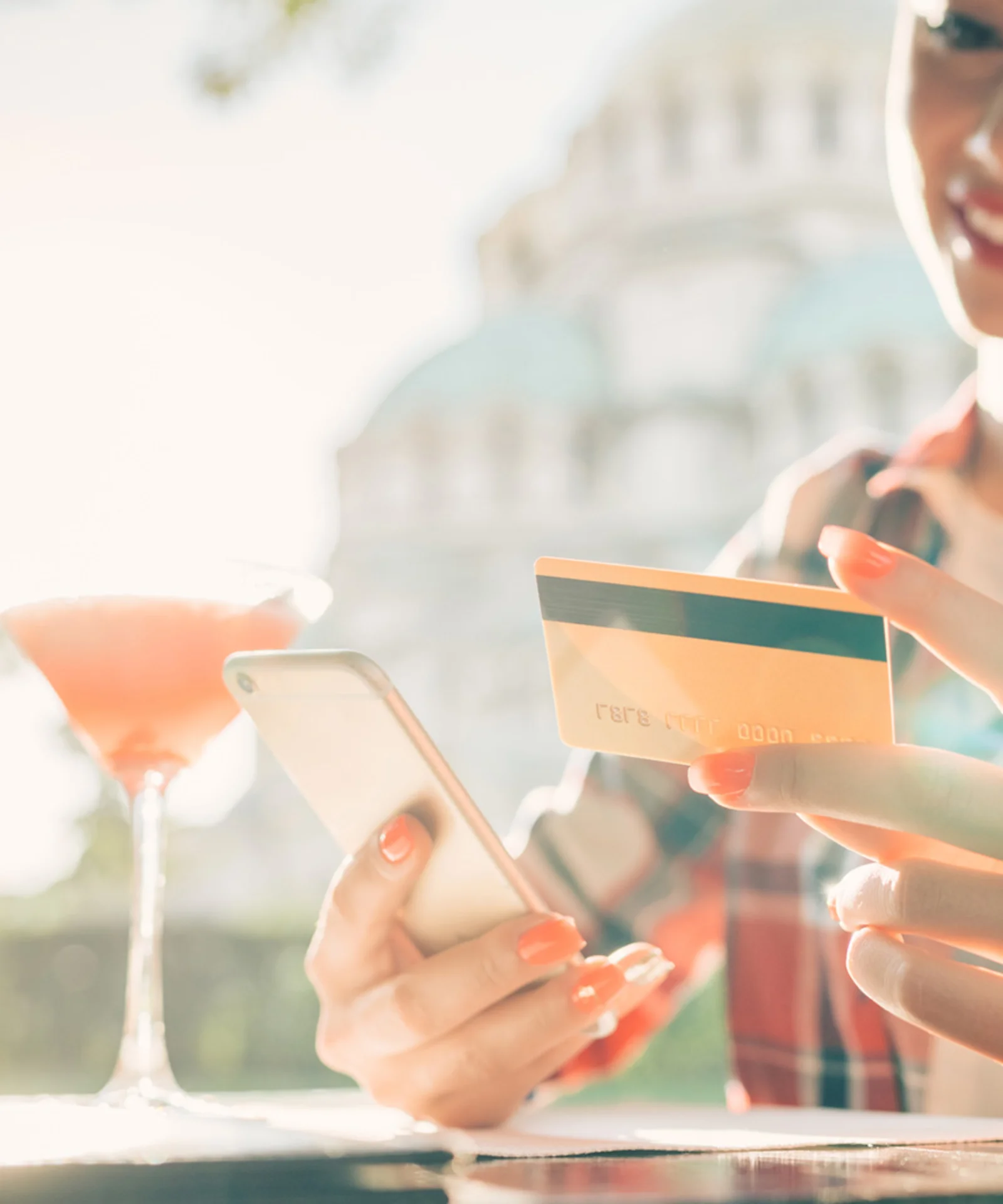 Una mujer sonriente sosteniendo un teléfono inteligente y una tarjeta de crédito, disfrutando de una bebida al aire libre, simbolizando la conveniencia y la facilidad de la banca personal moderna.