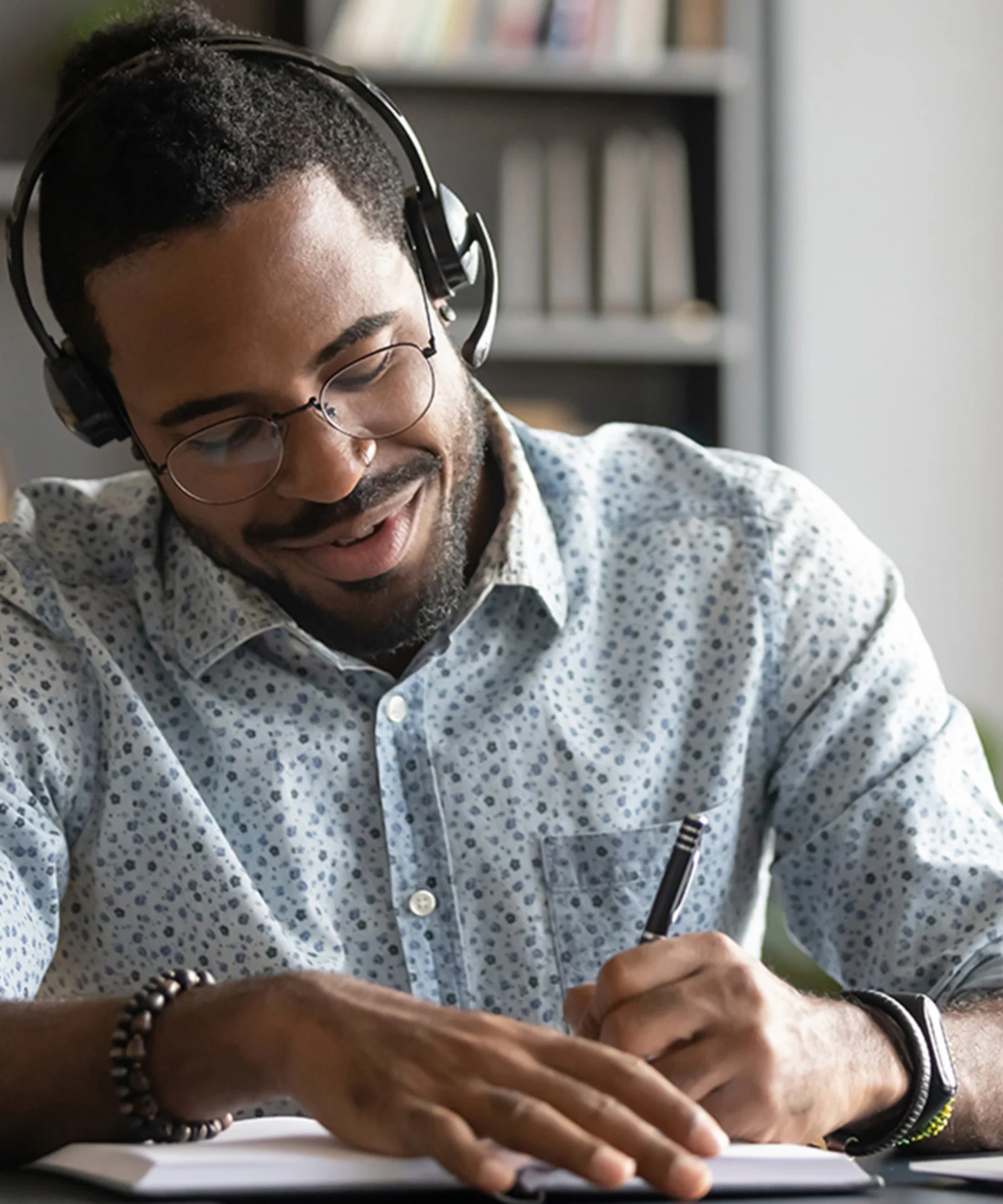 A man with headphones taking notes while listening to a podcast on his laptop, representing GFT&#039;s podcast library. He is seated in a cozy home office with bookshelves in the background, showcasing a comfortable and productive environment for learning and engagement.