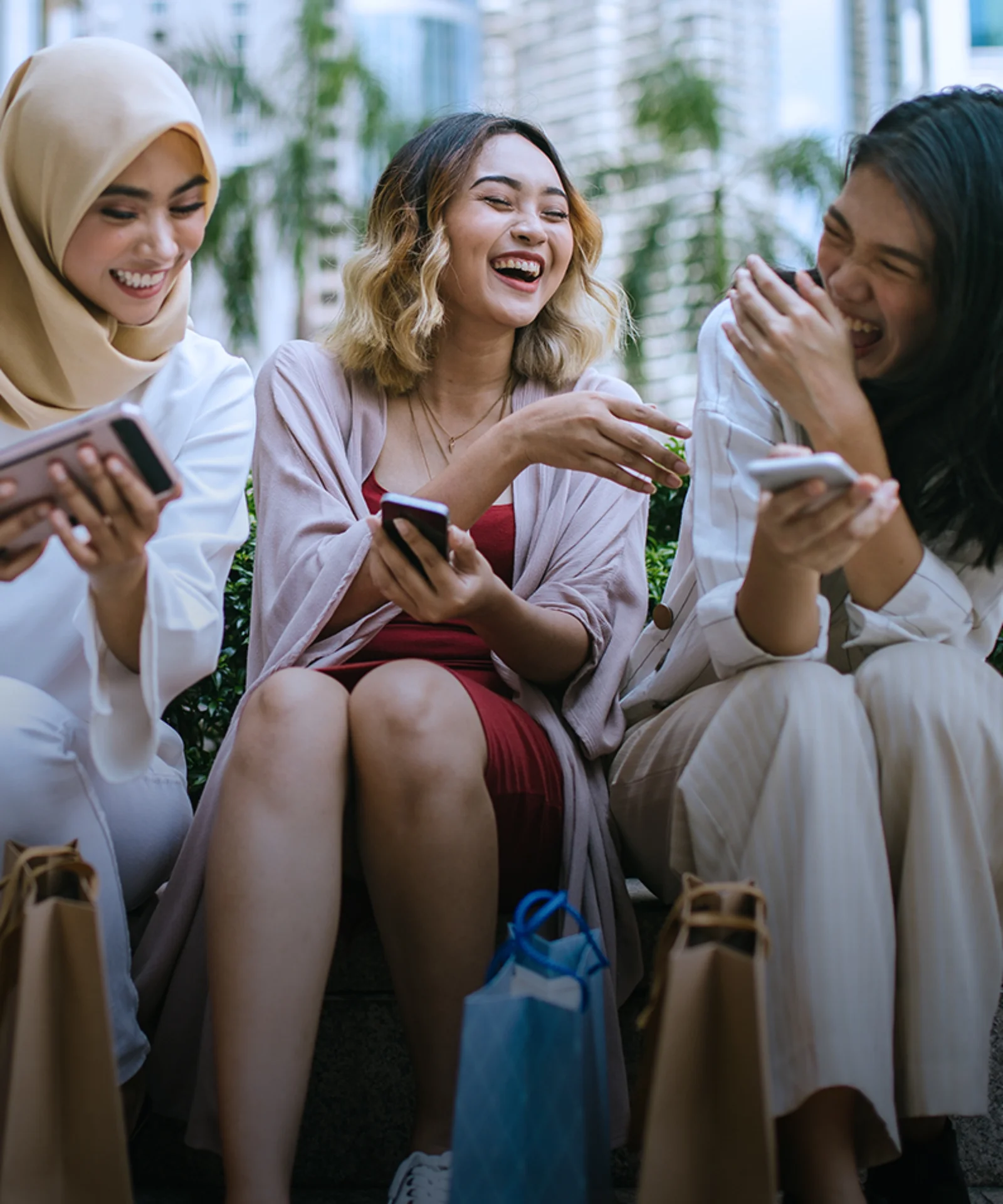 Three women sitting outdoors, laughing and sharing moments on their smartphones after a shopping trip, with shopping bags around them, symbolizing friendship and modern urban lifestyle.