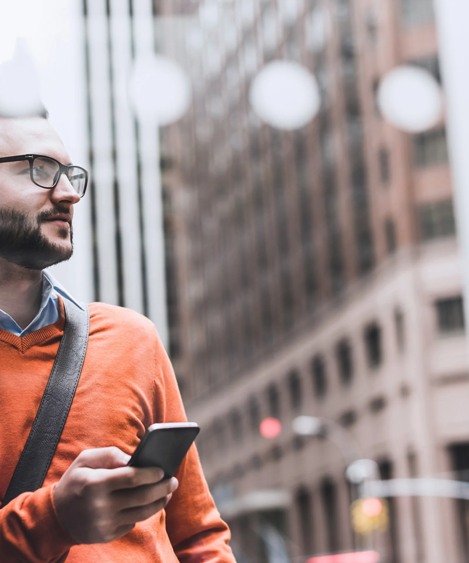 Man in an orange sweater holding a smartphone, standing in an urban environment.