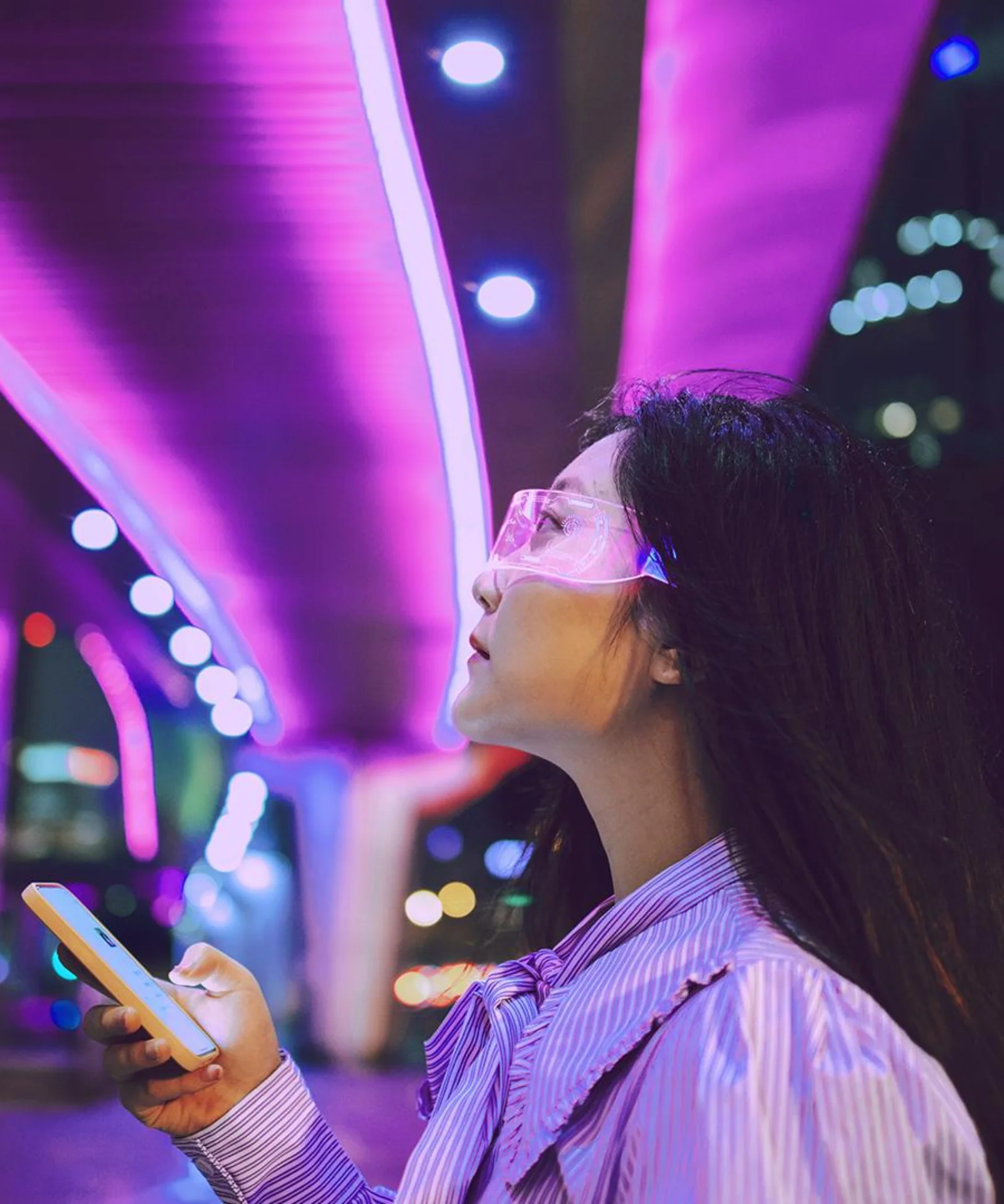 A woman wearing futuristic glasses and holding a smartphone, illuminated by purple city lights, representing the integration of AI technology into modern life.