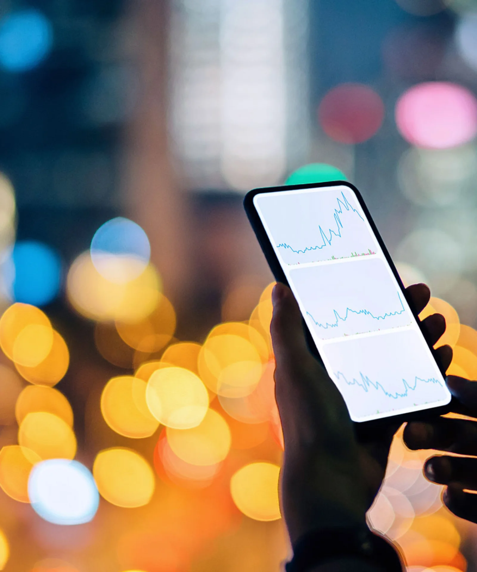 A person holding a smartphone displaying financial graphs, with a cityscape illuminated by vibrant lights in the background.