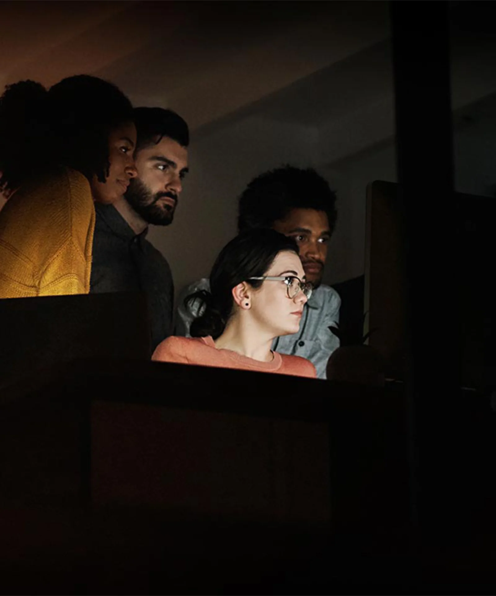 A focused team of four professionals working together on a project late at night, illuminated by the glow of computer screens in a dimly lit office.