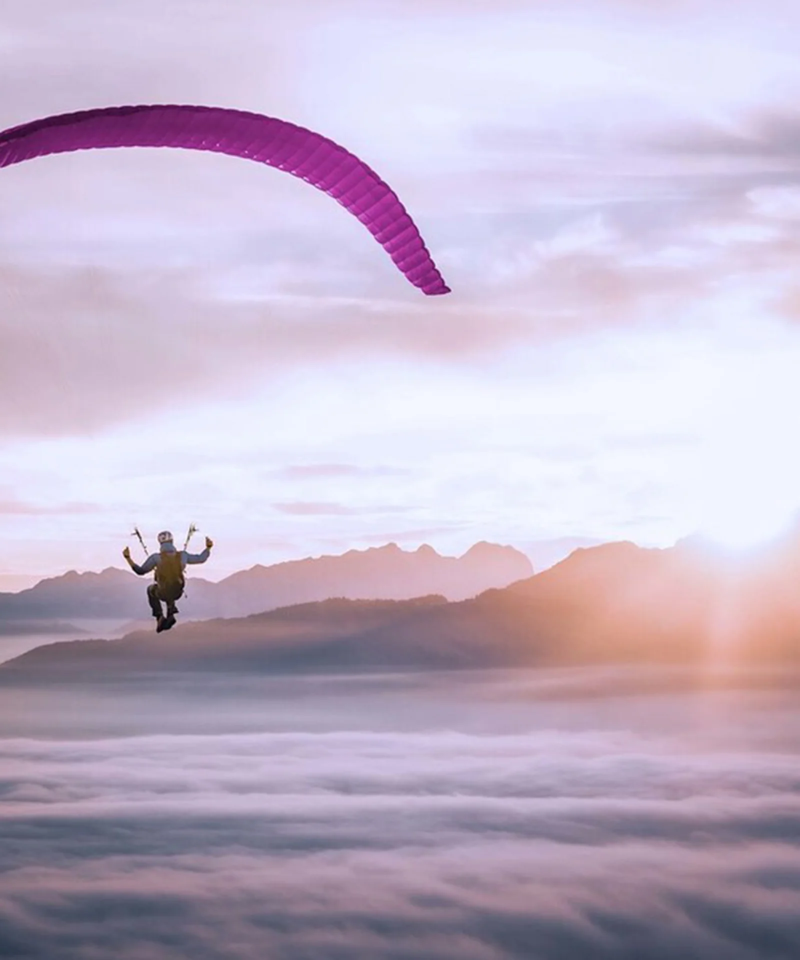 Esta imagem de tirar o fôlego captura a essência da liberdade e da aventura enquanto um parapente solitário voa graciosamente acima de um manto de nuvens. O parapente, com um dossel roxo vibrante, está posicionado contra o cenário deslumbrante de um nascer do sol, lançando um brilho dourado e quente sobre a cena. Os picos das montanhas distantes se elevam acima da camada de nuvens, adicionando profundidade e uma sensação de escala à imagem. A atmosfera serena e inspiradora destaca a beleza do parapente e a perspectiva única que ele oferece sobre o mundo. Este visual encapsula perfeitamente a emoção e a tranquilidade de voar em céu aberto.