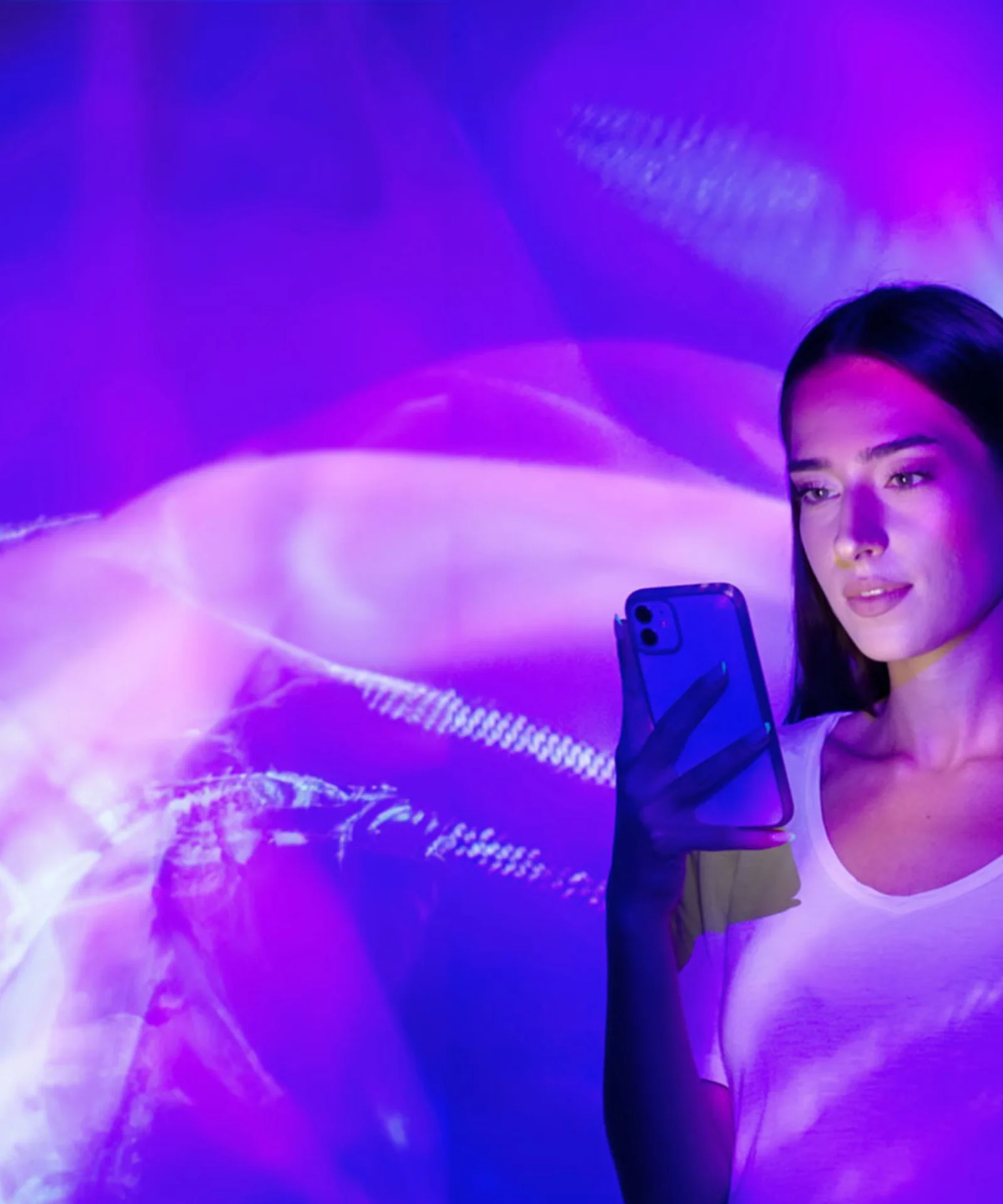 Une femme tenant un smartphone se tient devant un écran lumineux violet et rose vibrant et abstrait.