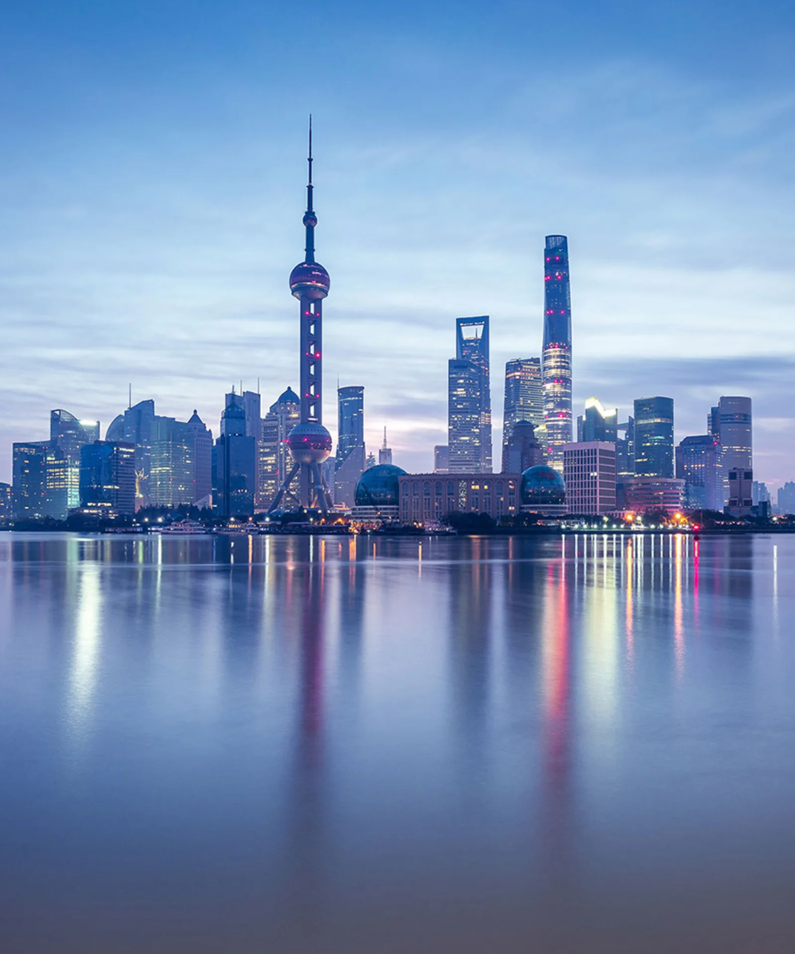 A panoramic view of Shanghai’s skyline at dusk, featuring modern skyscrapers and the iconic Oriental Pearl Tower, reflecting in the calm waters of the Huangpu River.