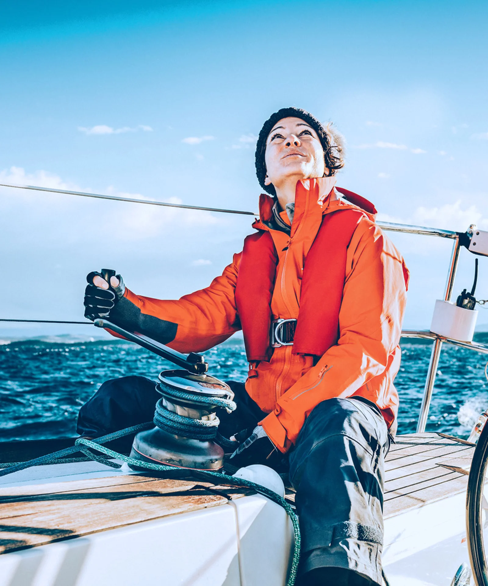 Person in bright orange sailing gear steering a yacht on open waters under a clear blue sky.