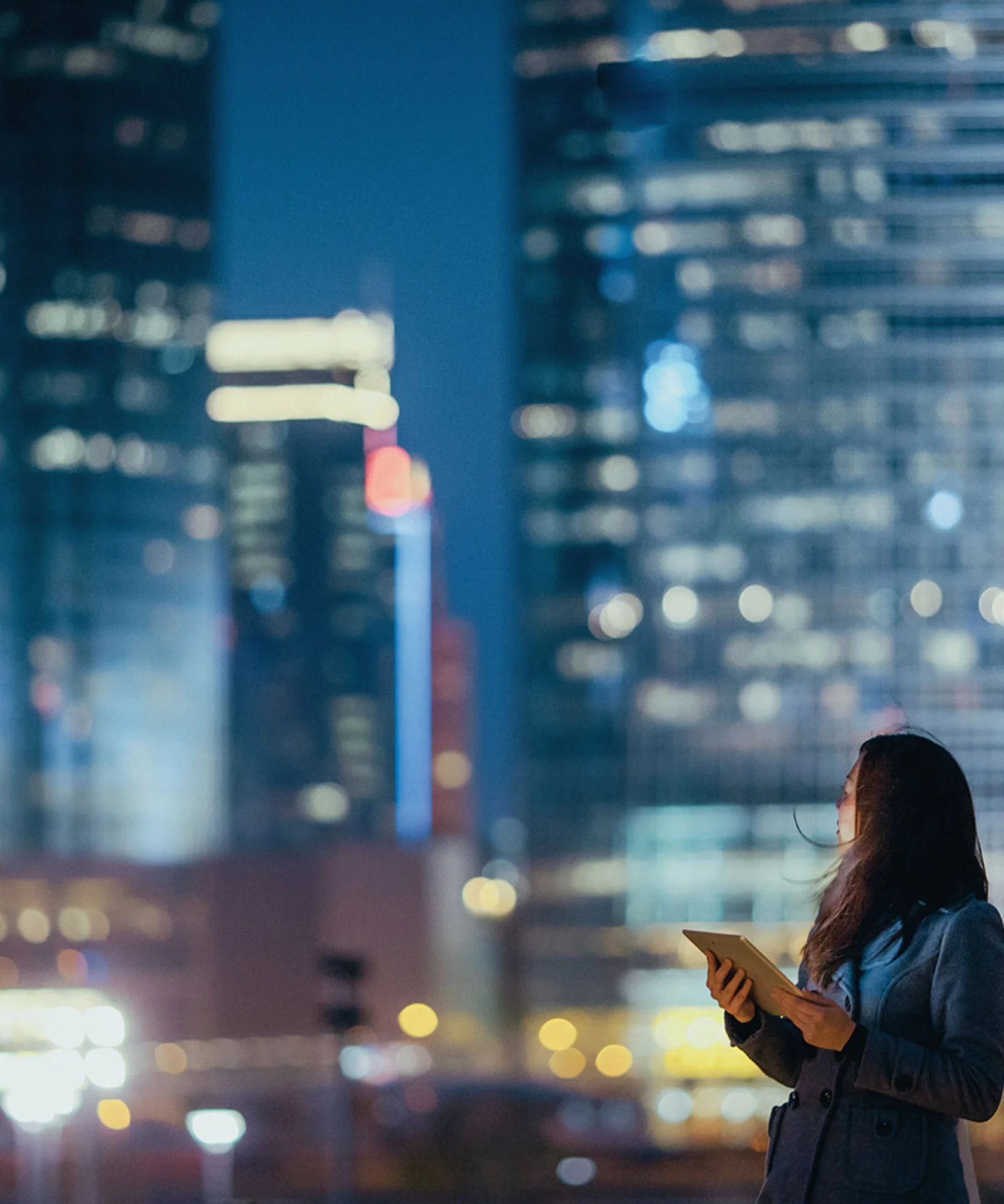 A woman holding a tablet looks up at illuminated skyscrapers in a city at night, symbolizing digital innovation and technology in an urban environment.