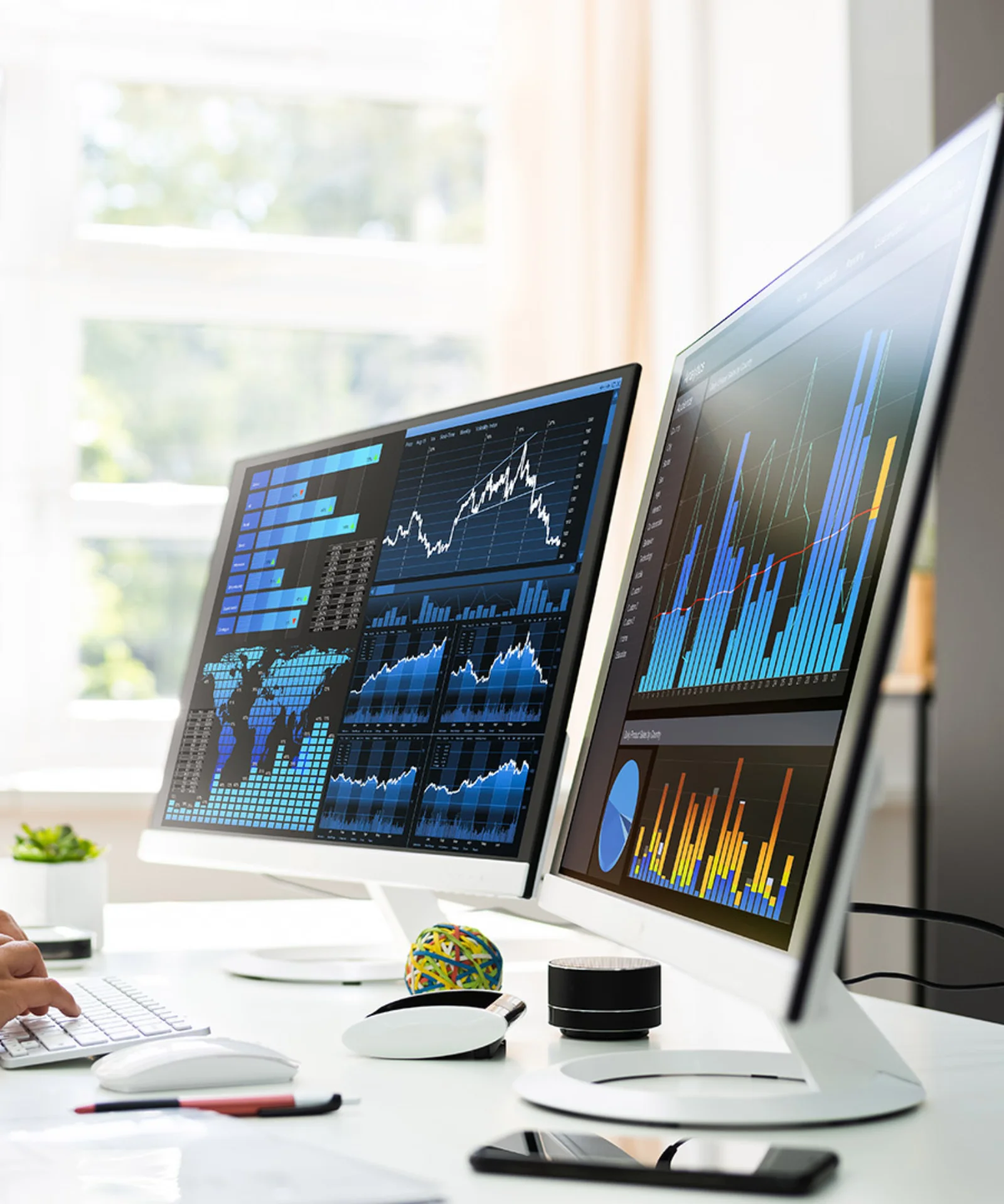 Woman in a white shirt working on a computer with dual monitors displaying various data charts and graphs in a bright office.