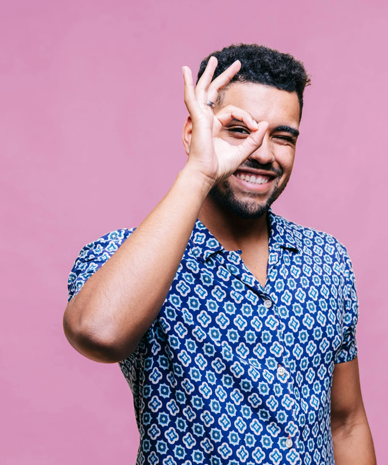 A cheerful young man making an &#039;OK&#039; sign with his hand against a pink background.