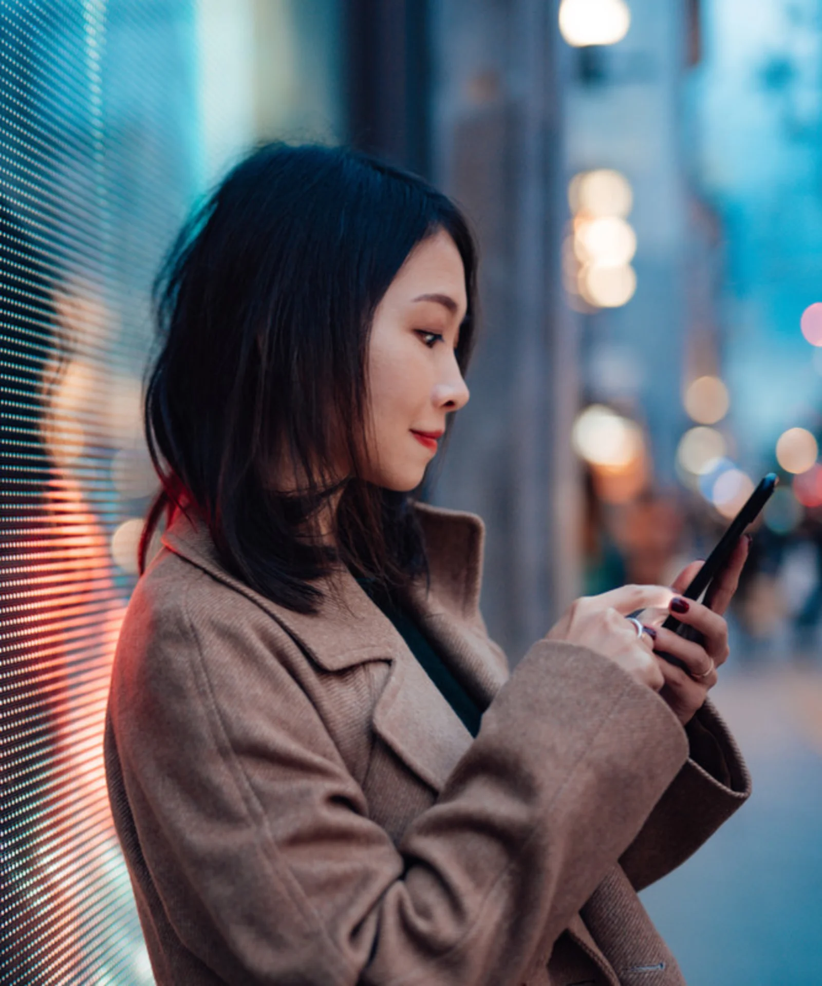 Una mujer de pie frente a una pantalla digital por la noche, usando su teléfono inteligente. La luz de la pantalla se refleja en su rostro y en el área circundante, creando un ambiente moderno y tecnológico.