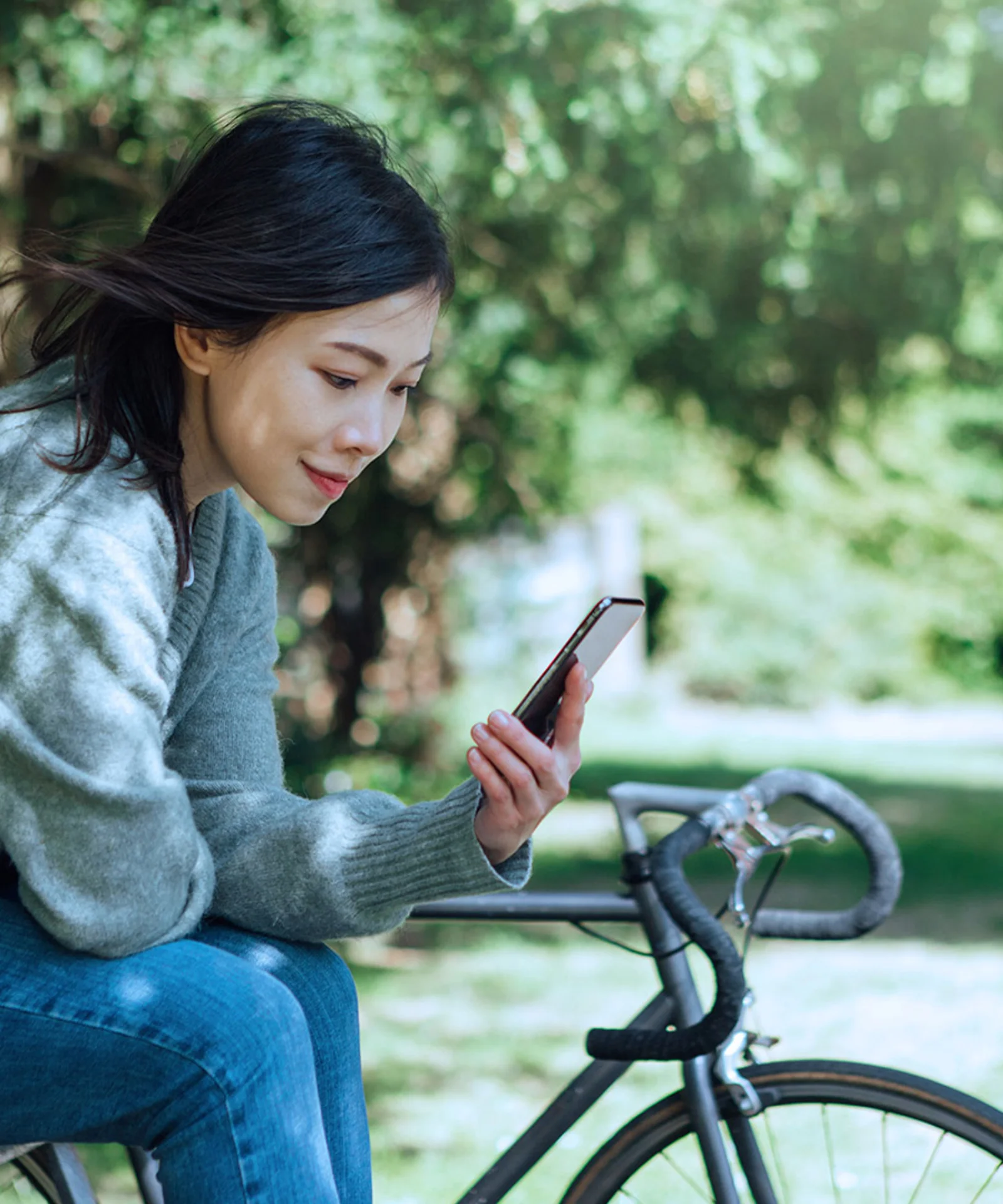 A woman sitting on a bicycle, looking at her phone, in a green park setting, representing sustainability by design