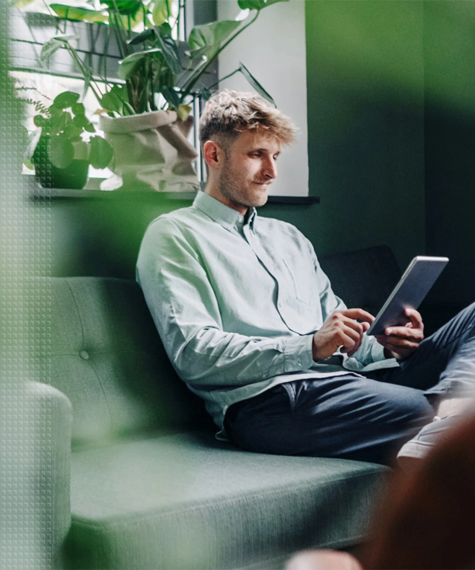 A person sitting on a couch in a green, plant-filled room, using a tablet, symbolising eco-friendly technology practices.