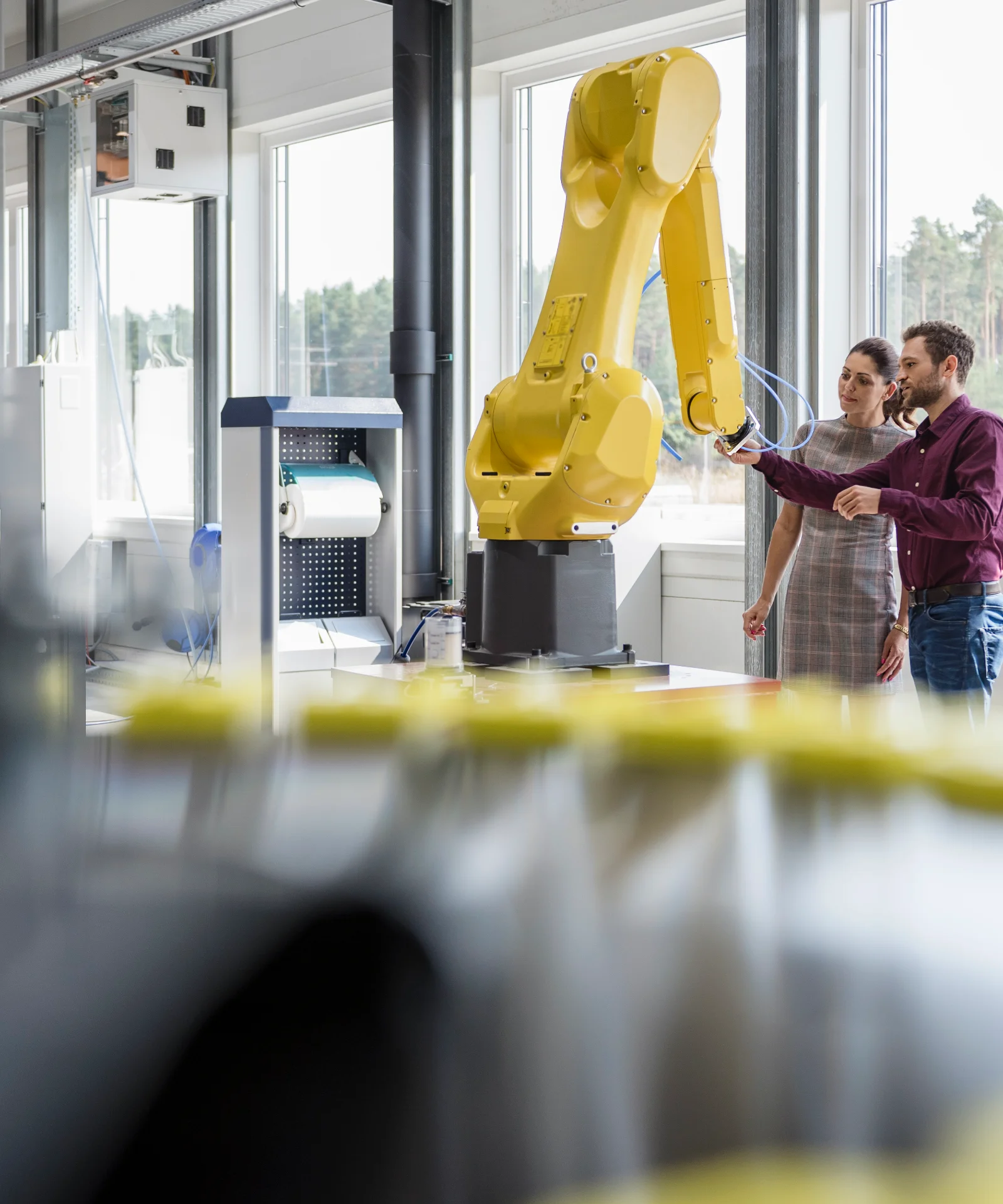 Mann und Frau betrachten einen Roboterarm in einer Fabrikhalle, im Hintergrund ist ein Wald zu sehen. Key Visual für Industry Solutions.