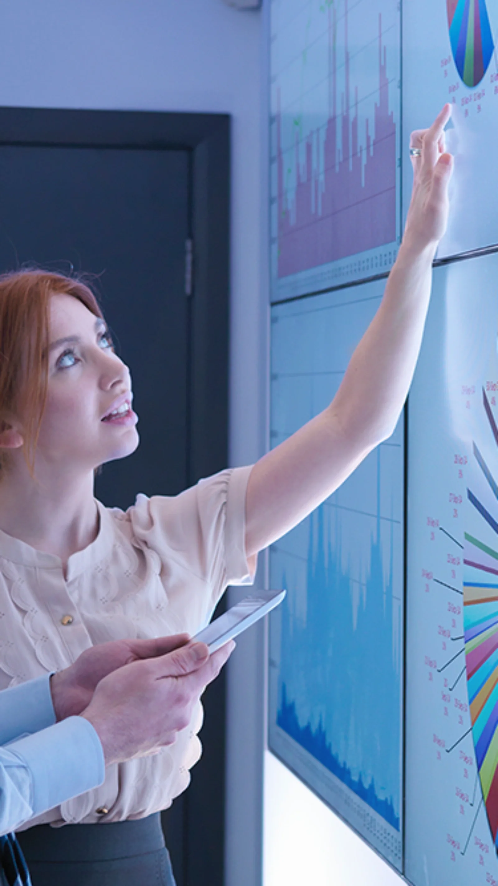 Two business analysts examining large digital screens displaying complex data visualizations, graphs, and charts, focused on analyzing trends.