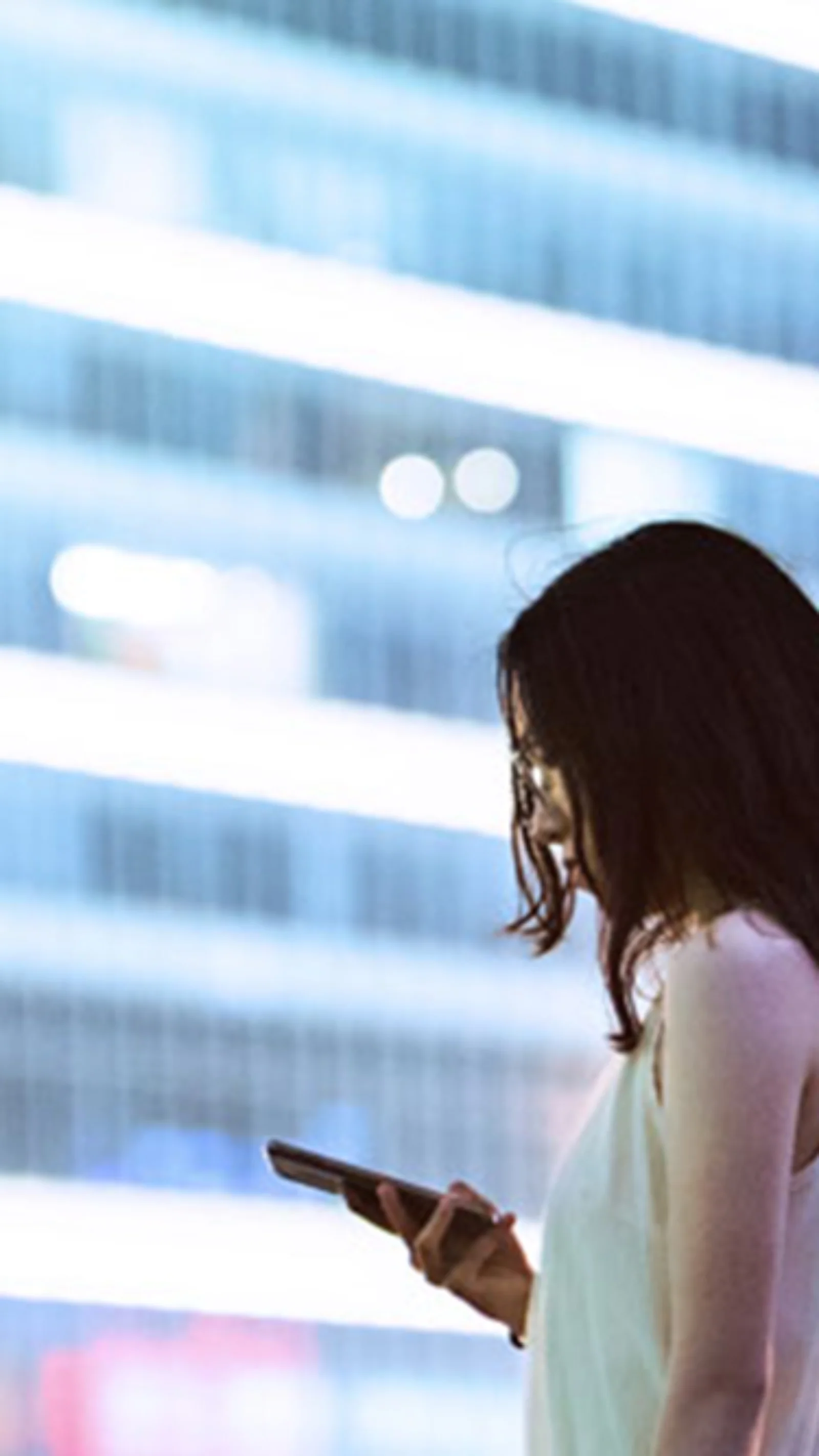 A professional woman using her smartphone with a modern cityscape in the background, symbolizing the impact of Central Bank Digital Currencies (CBDCs) on the future of commercial banking.