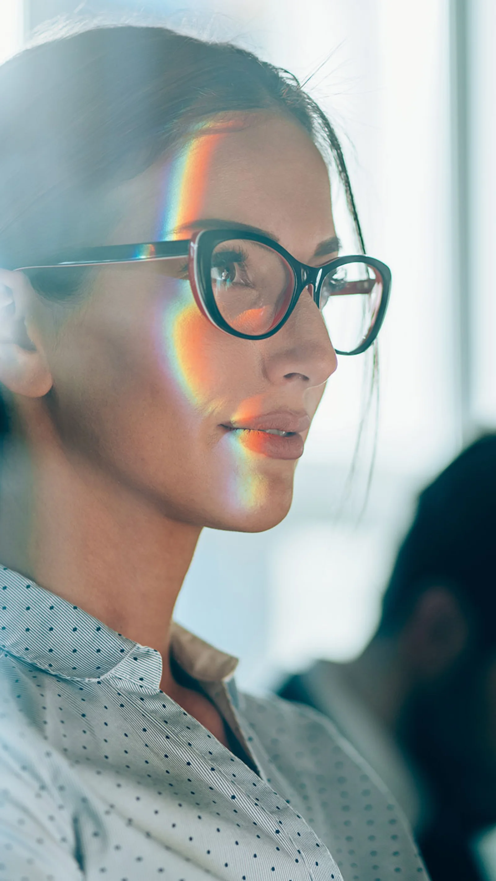 A confident woman wearing glasses looks ahead with a focused expression, with a subtle rainbow reflection on her face, symbolising strategic thinking and modern business acumen.