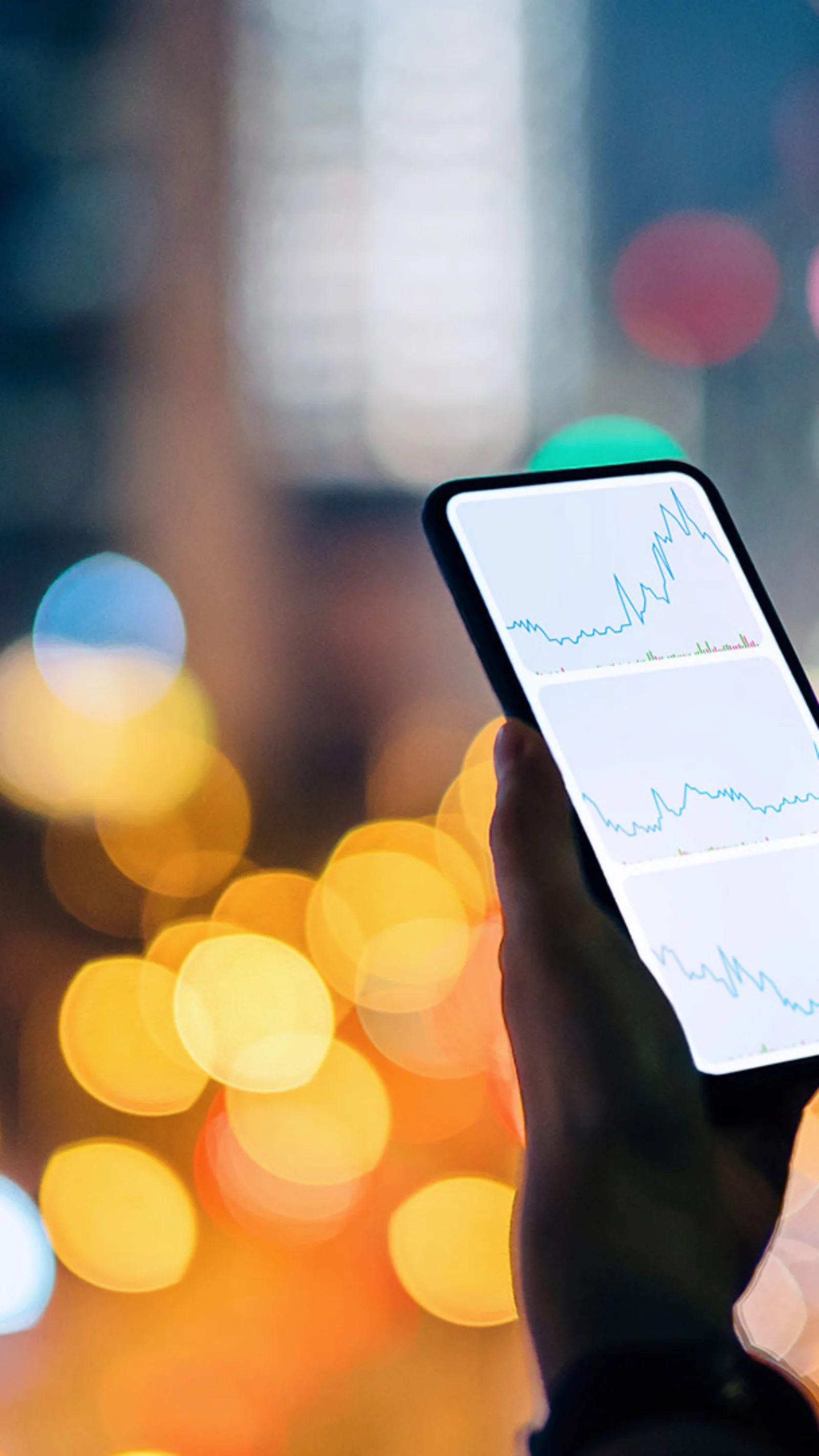 A person holding a smartphone displaying financial graphs, with a cityscape illuminated by vibrant lights in the background.