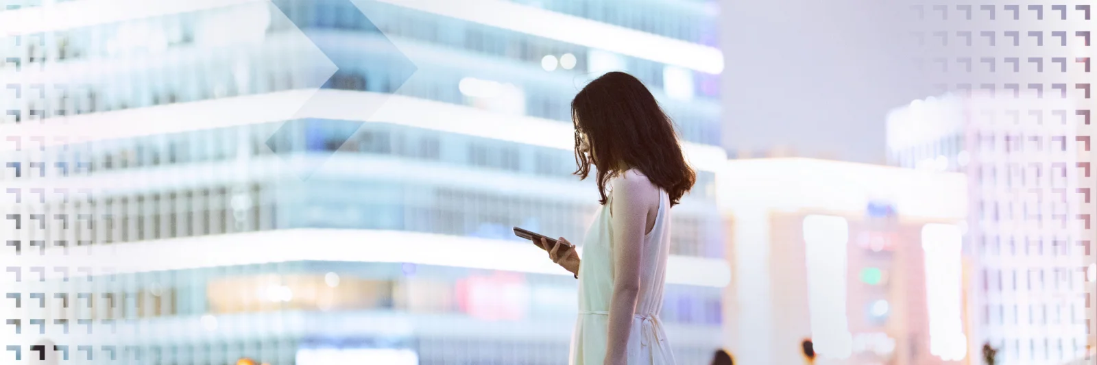 Eine Frau im weißen Kleid steht in einer modernen Stadt und schaut auf ihr Smartphone, umgeben von beleuchteten Gebäuden.