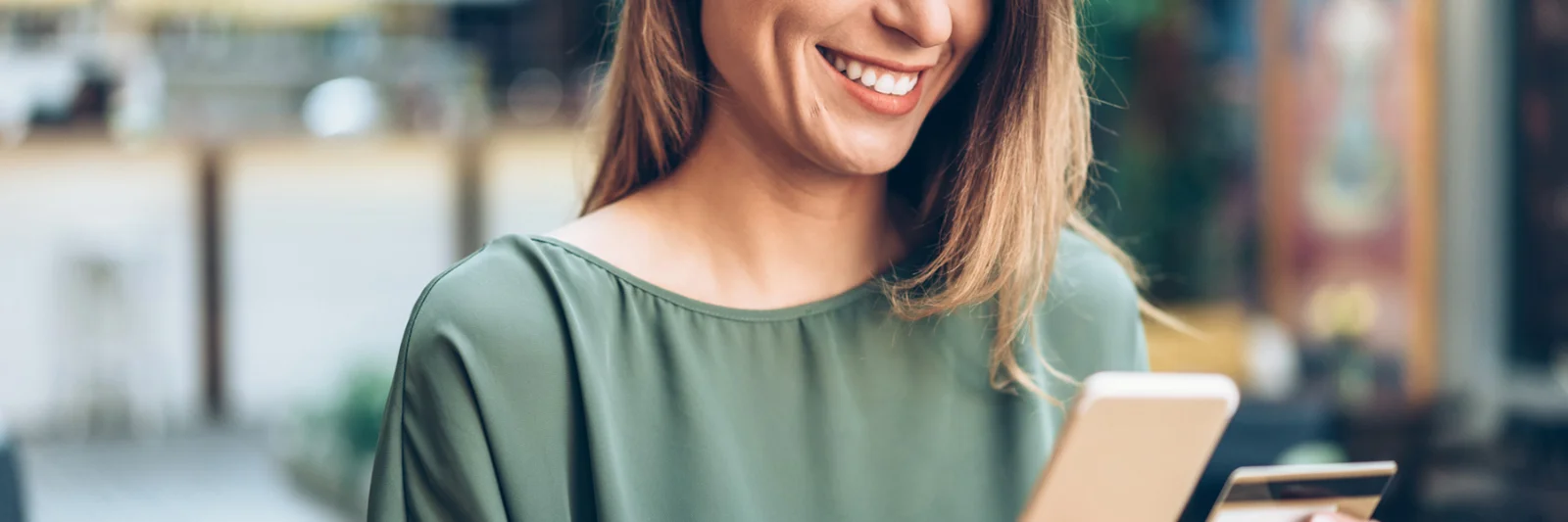 Una mujer sonriente usa un teléfono inteligente y sostiene una tarjeta de crédito, lo que representa la conveniencia de una nueva aplicación móvil de banca comercial.