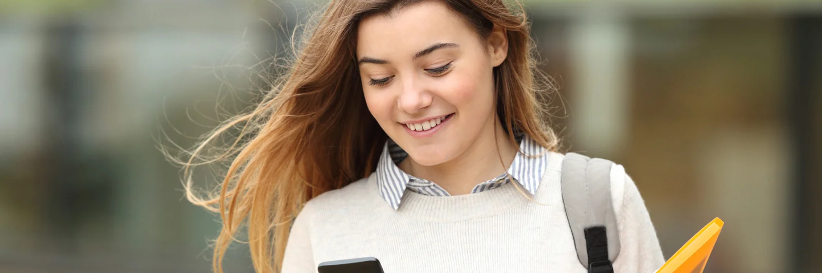 A young woman smiling while using her smartphone, holding a folder, symbolising the convenience and accessibility of the Crédit Agricole BankMeApp.