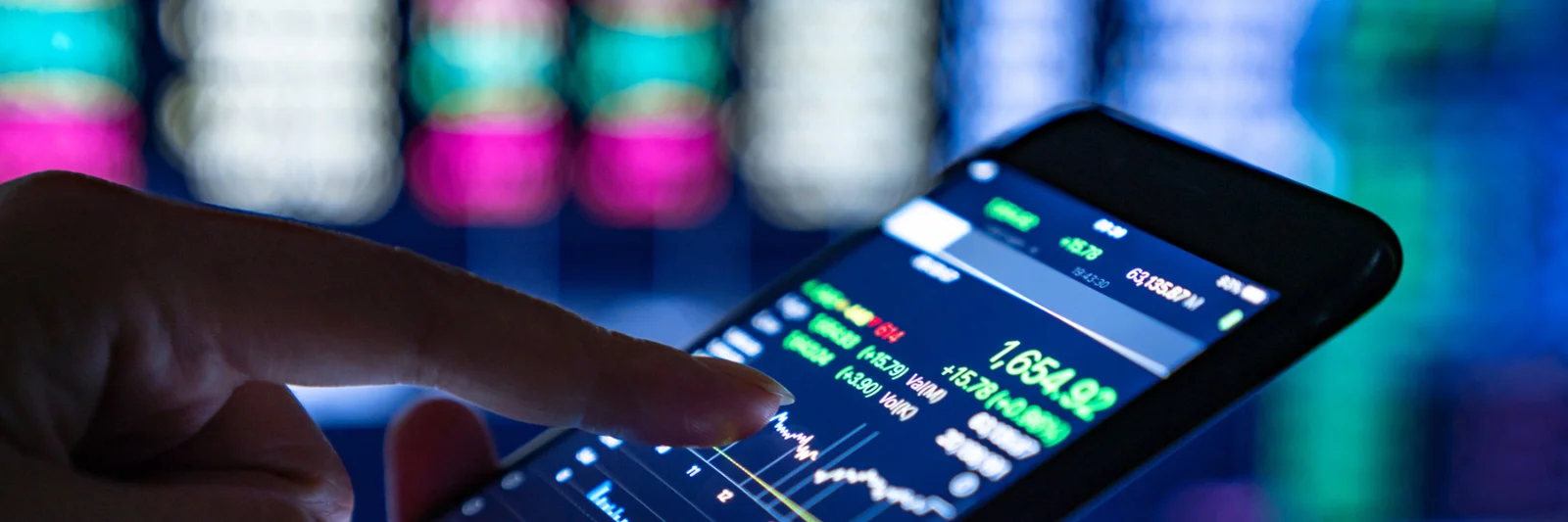 A close-up of a hand using a smartphone to monitor stock market data with blurred financial screens in the background.