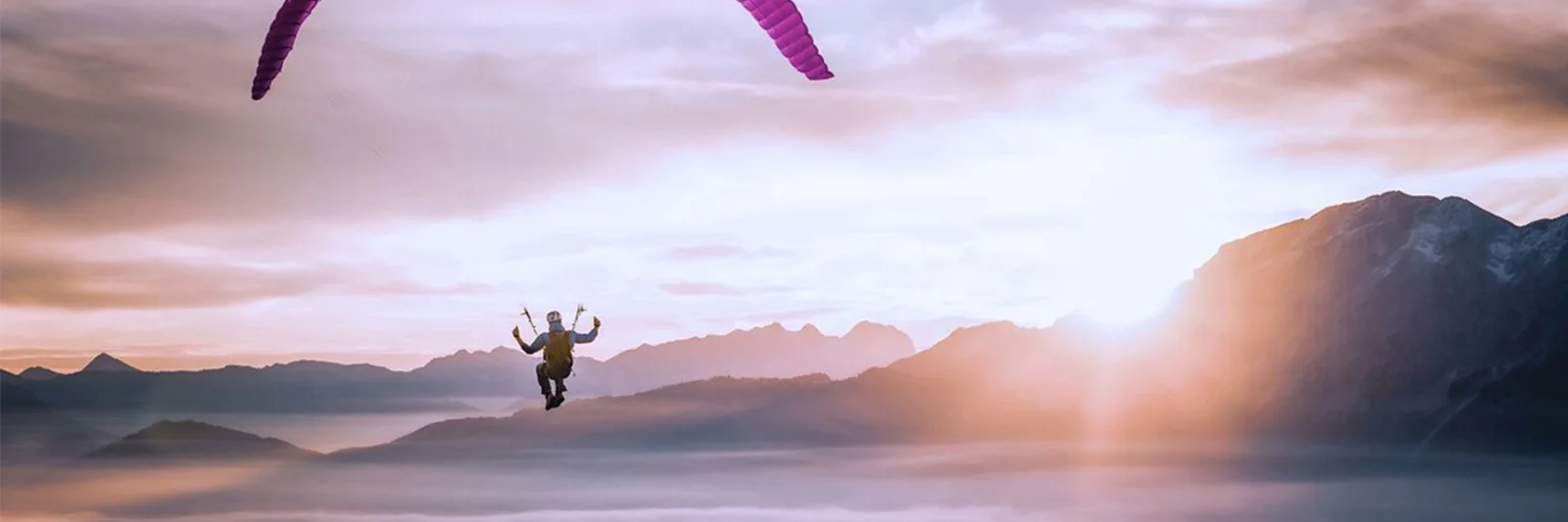 A paraglider silhouetted against a majestic sunrise, floating above a sea of clouds with mountain peaks in the background.