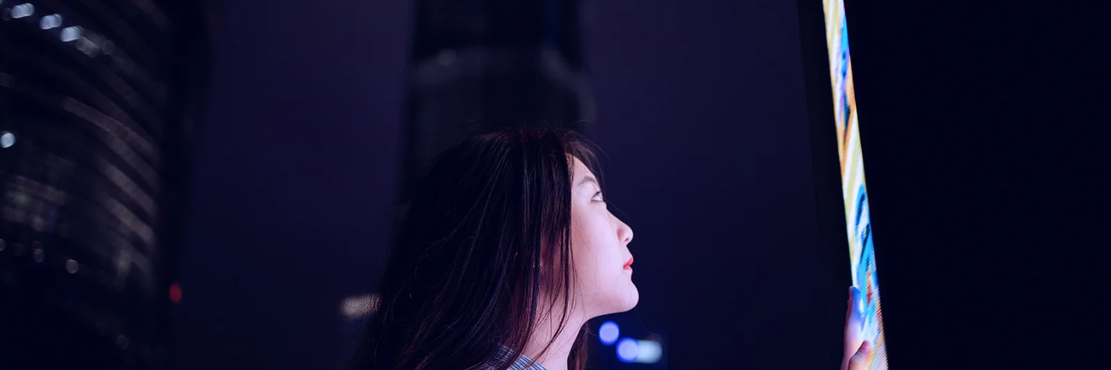 A woman interacts with a large, illuminated screen at night, set against the backdrop of a cityscape with towering buildings.