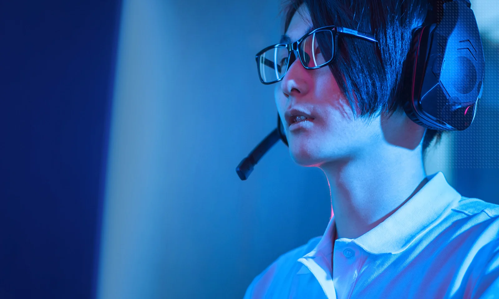 A focused gamer with glasses and a headset is immersed in a game, illuminated by blue lighting.