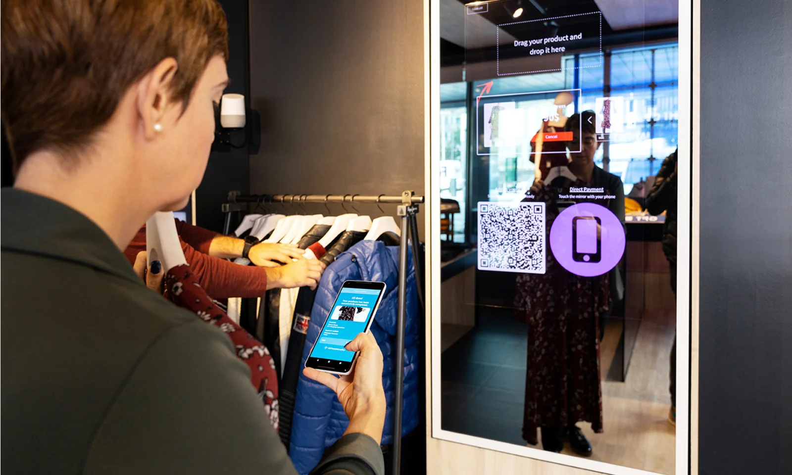 A participant using a smartphone to interact with a smart mirror display in a design product innovation workshop. The smart mirror shows product information, QR codes, and payment options, enhancing the retail shopping experience.