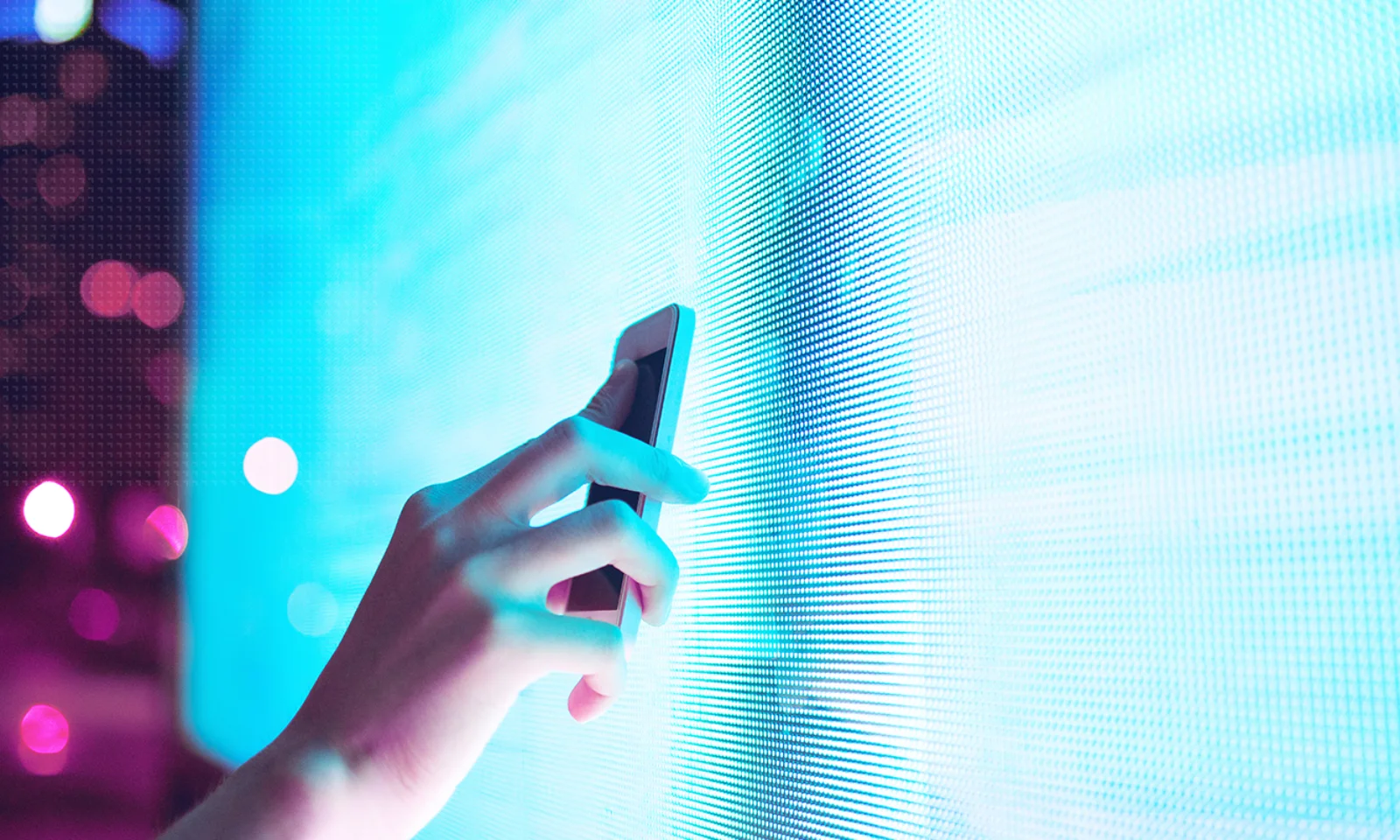 Close-up of a hand holding a smartphone near a bright, neon-blue digital display, highlighting the interaction between mobile technology and digital media.