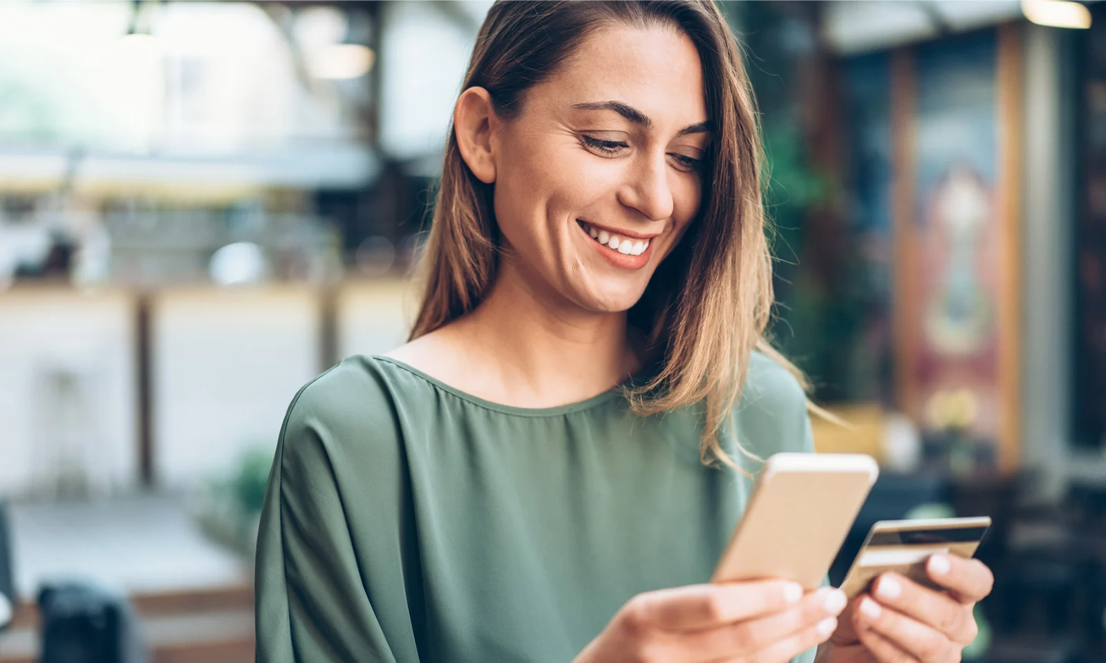 Smiling woman using a smartphone and holding a credit card for an online payment in a cafe.