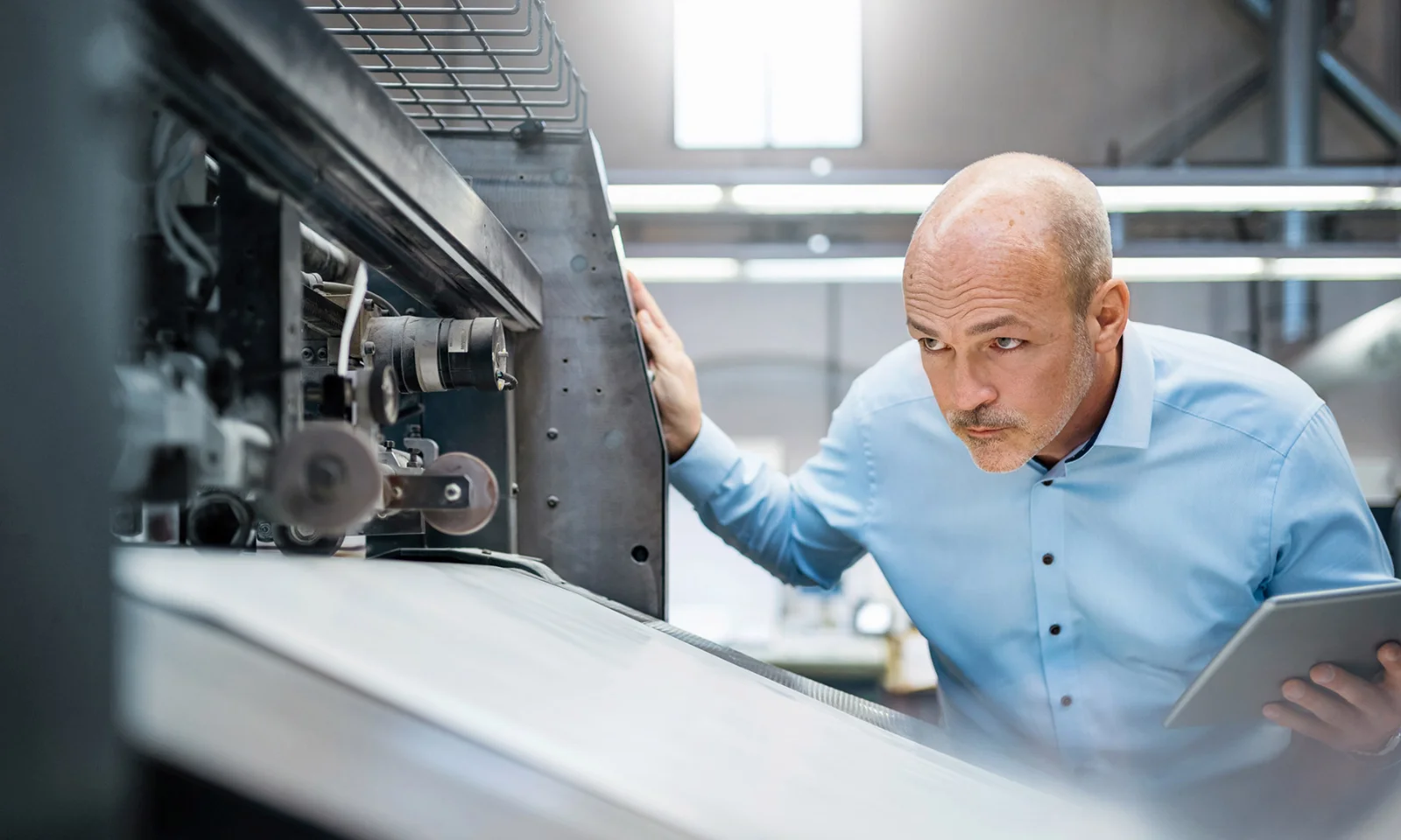 Engineer closely inspecting industrial machinery while holding a tablet in a manufacturing facility.