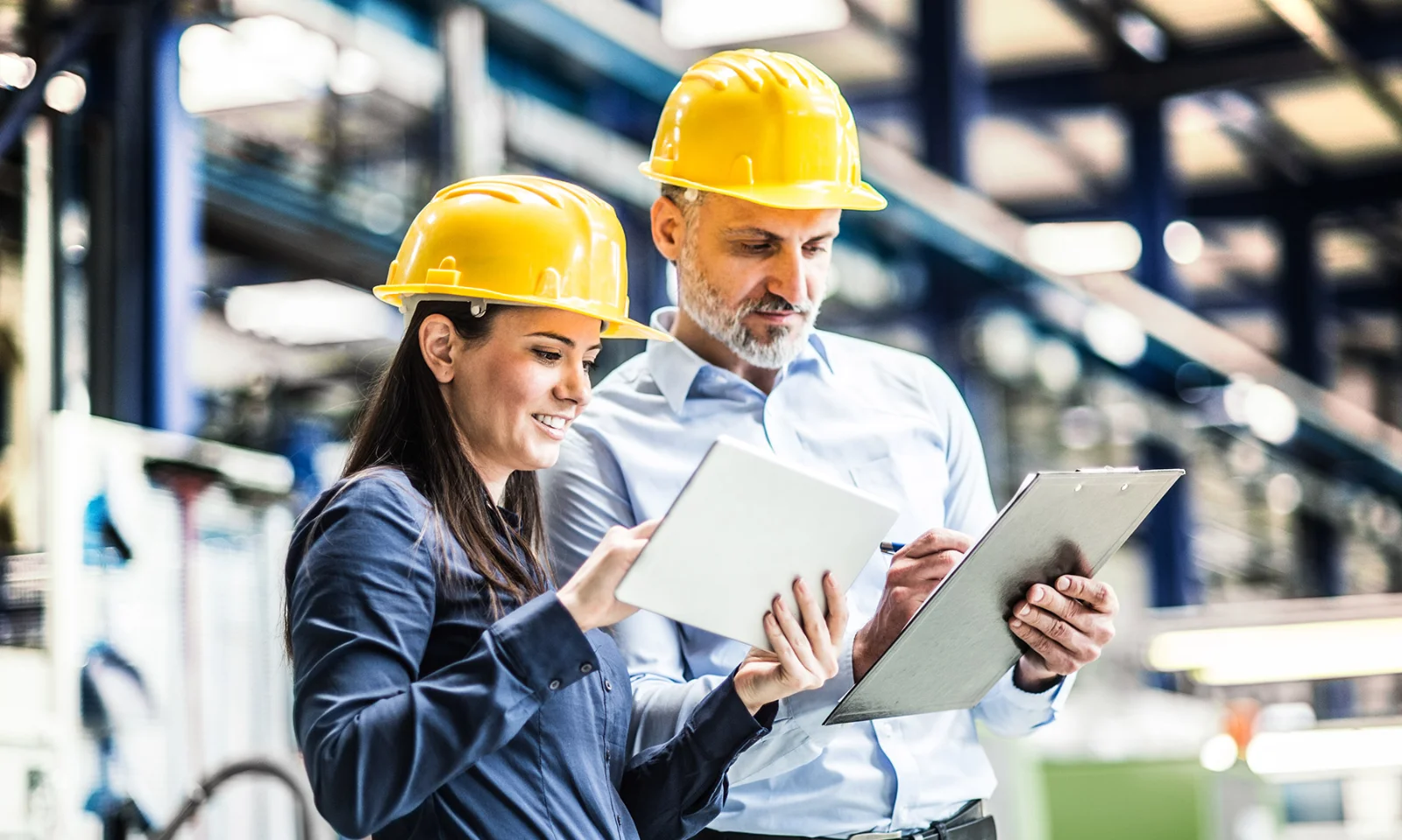 Two engineers in yellow hard hats reviewing production plans on a tablet and clipboard in an industrial facility.