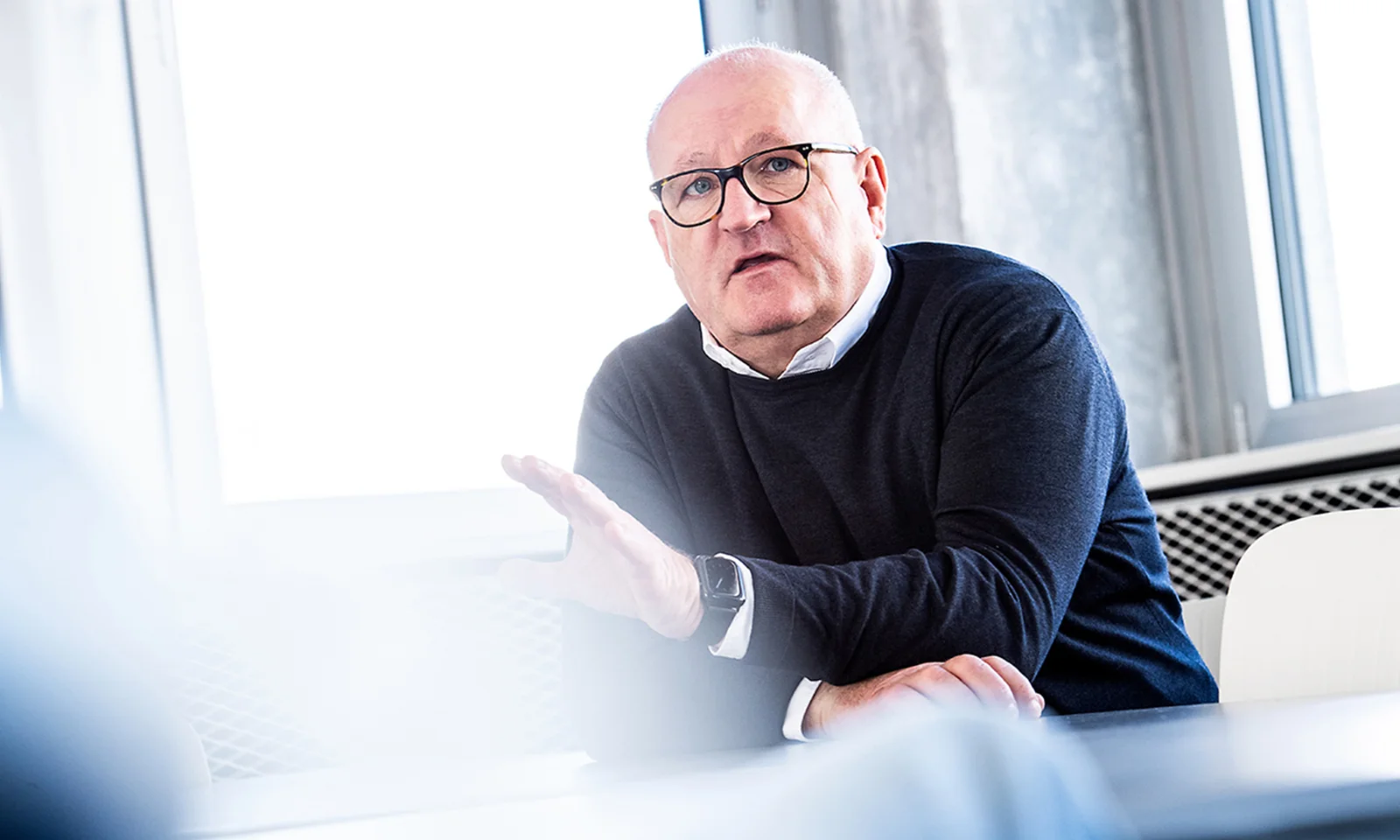 Ulrich Dietz actively engaging in a discussion, wearing glasses and a dark sweater, in a bright office setting