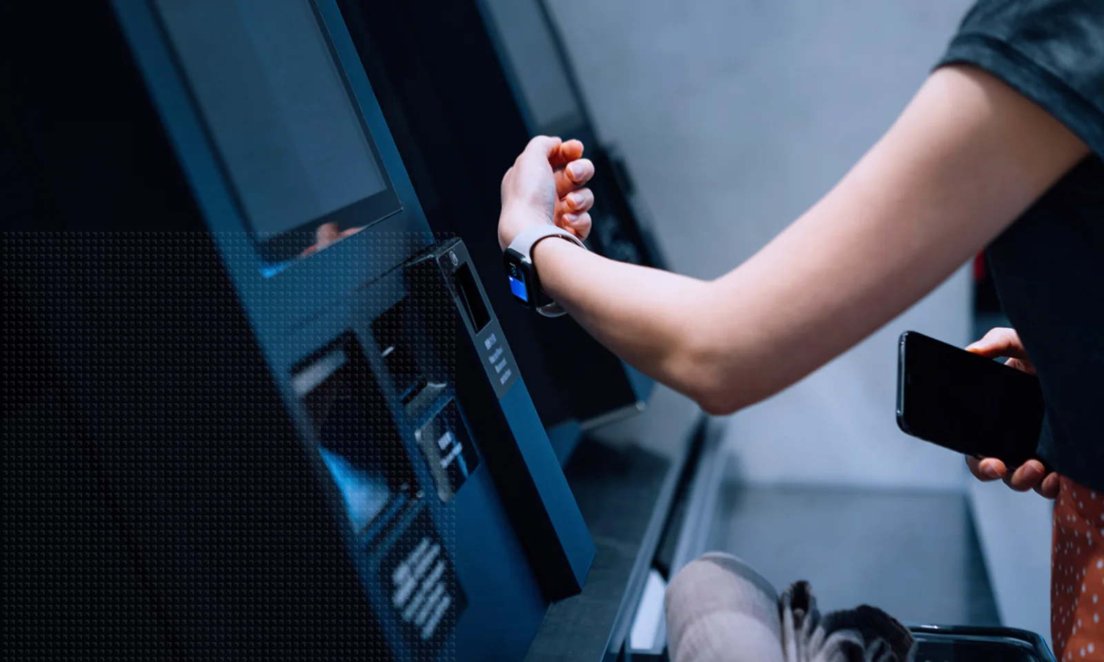 A person uses a smartwatch to make a contactless payment at a self-service kiosk while holding a smartphone and shopping basket.