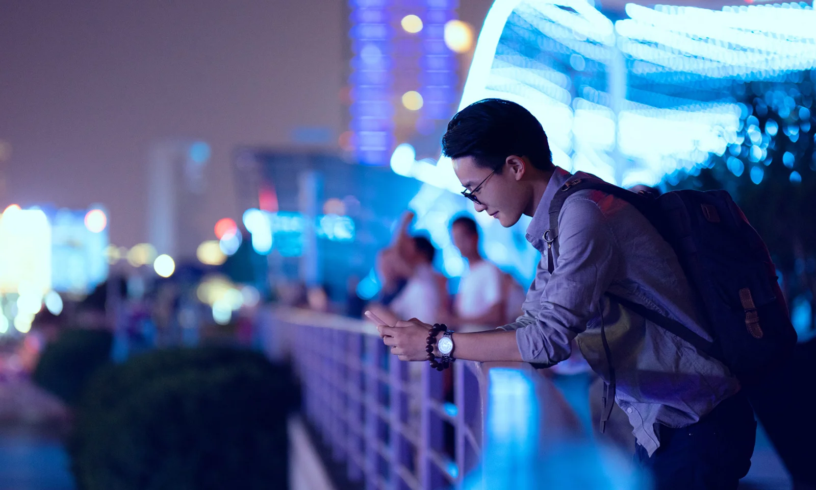 A young professional with a backpack using a mobile device while leaning on a railing in a vibrant, illuminated city at night.